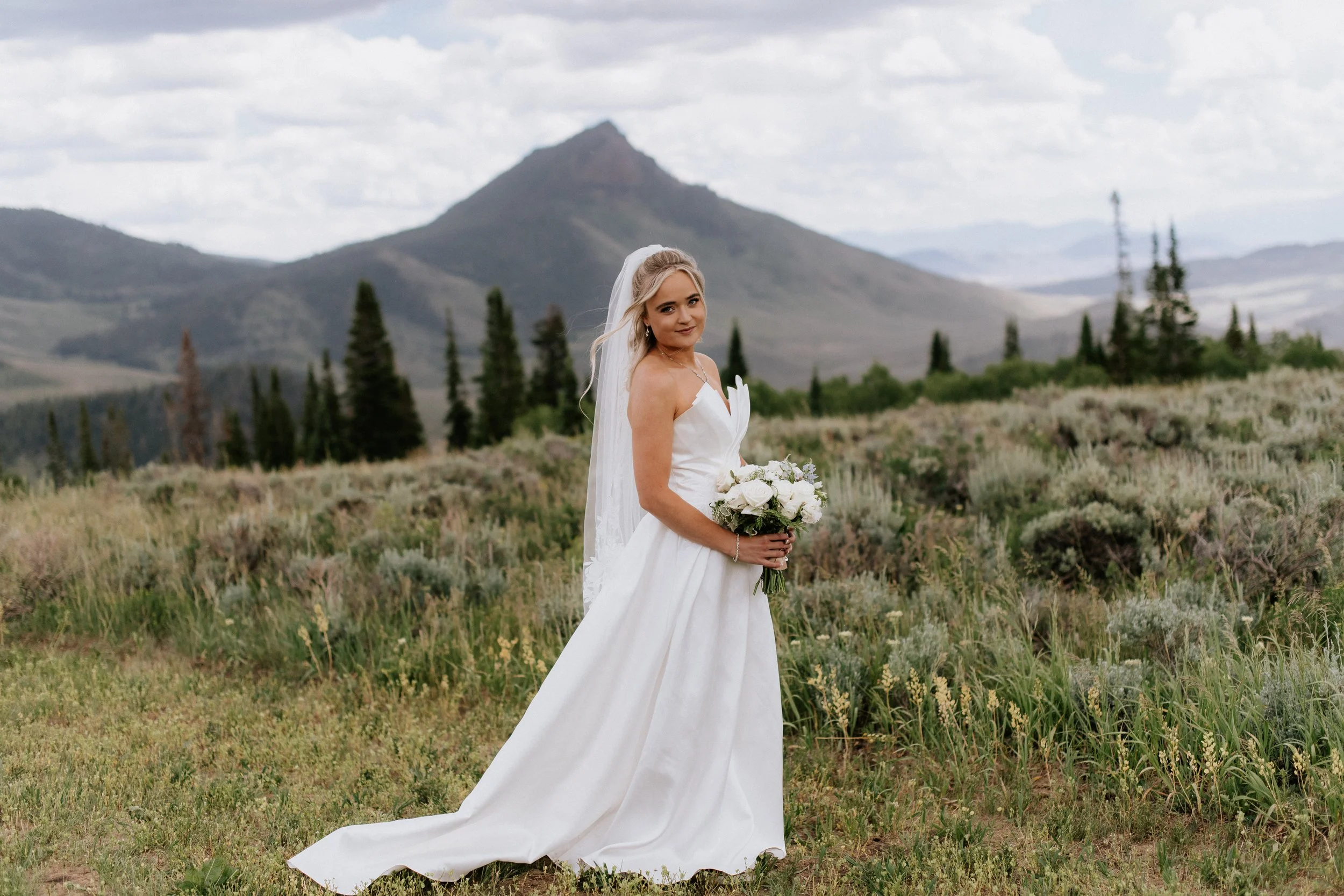 A bride in a white wedding dress with a veil holding a bouquet of white roses in a field with green vegetation. In the background, there are trees and a mountain under a partly cloudy sky.
