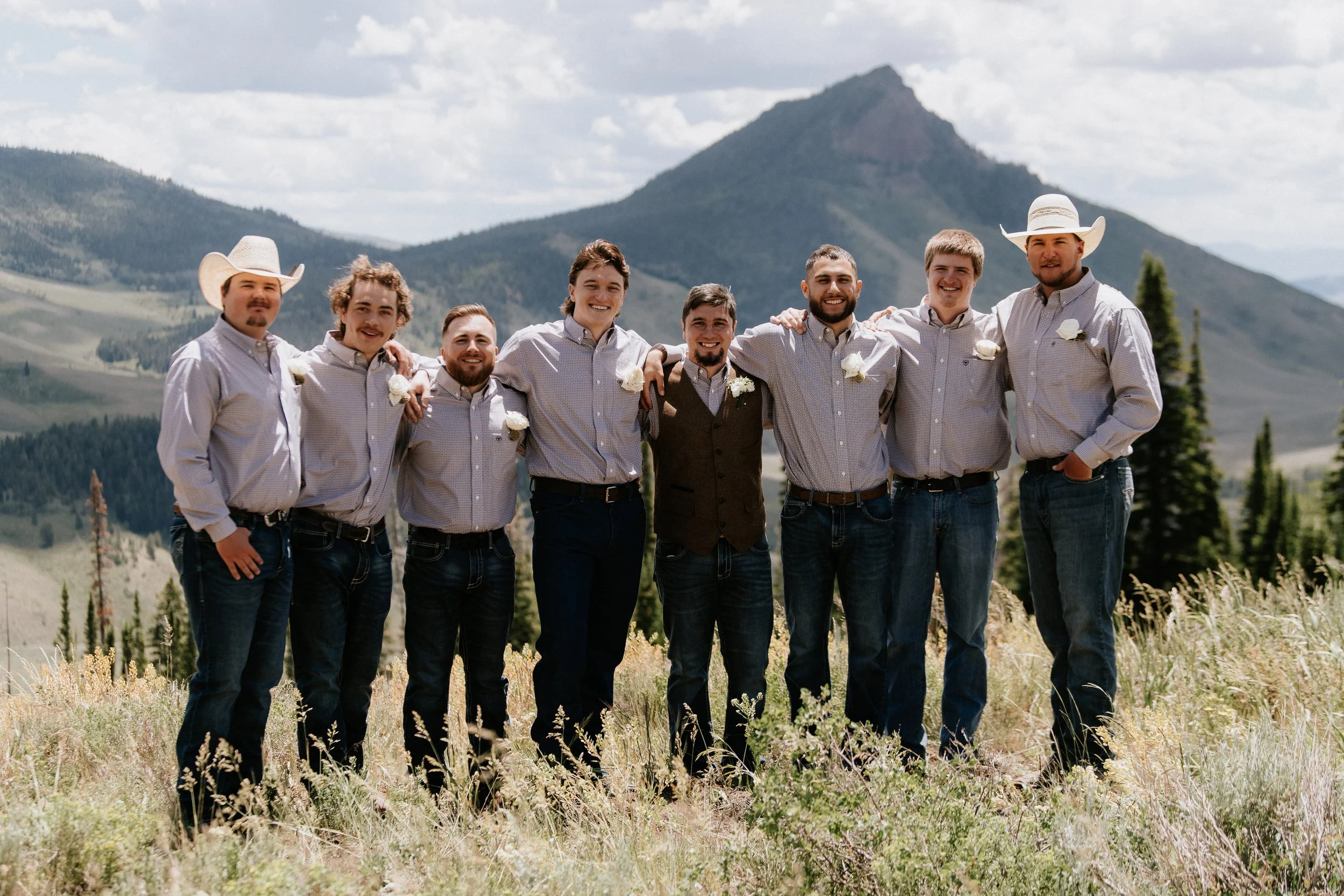 Group of eight men standing outdoors in a grassy field with mountains and cloudy sky in the background, smiling and arm-in-arm, dressed in casual shirts and jeans. Two men are wearing cowboy hats, and some have white flower boutonnières.