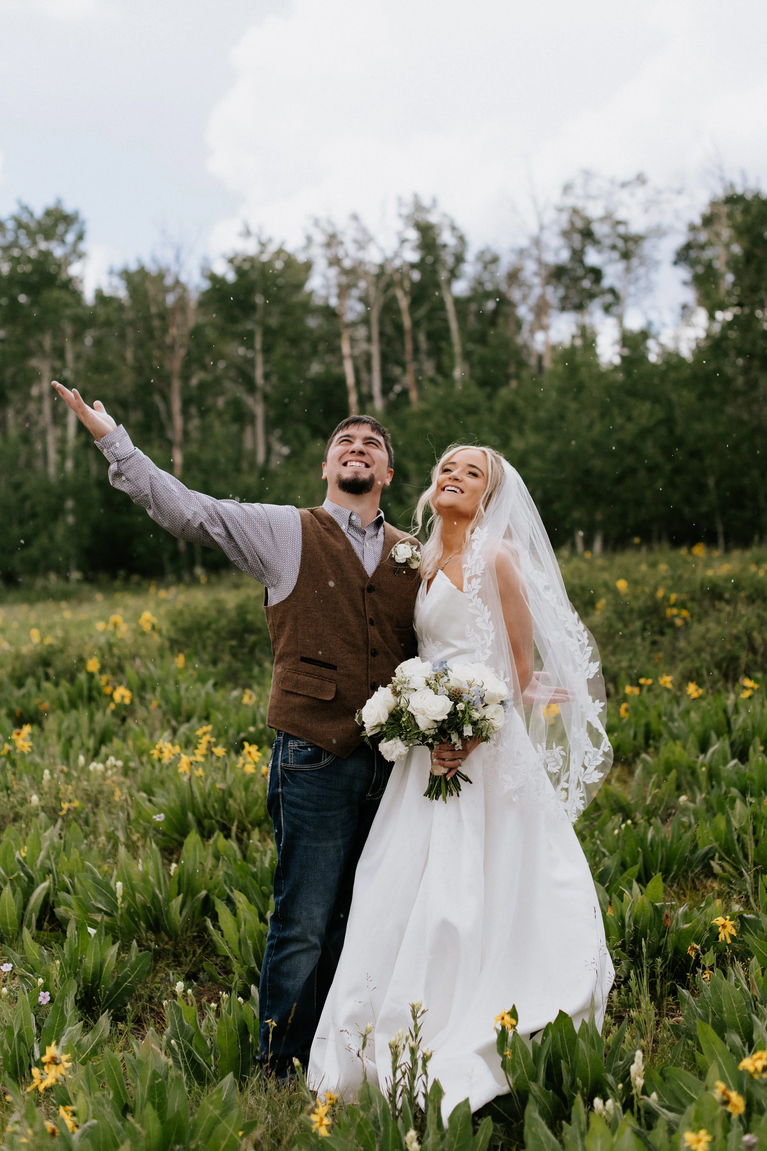 A newlywed couple standing in a field of yellow flowers with trees in the background. The groom is smiling and reaching up, while the bride holds a bouquet of white flowers and is smiling. The bride is wearing a white wedding dress and veil, and the 
