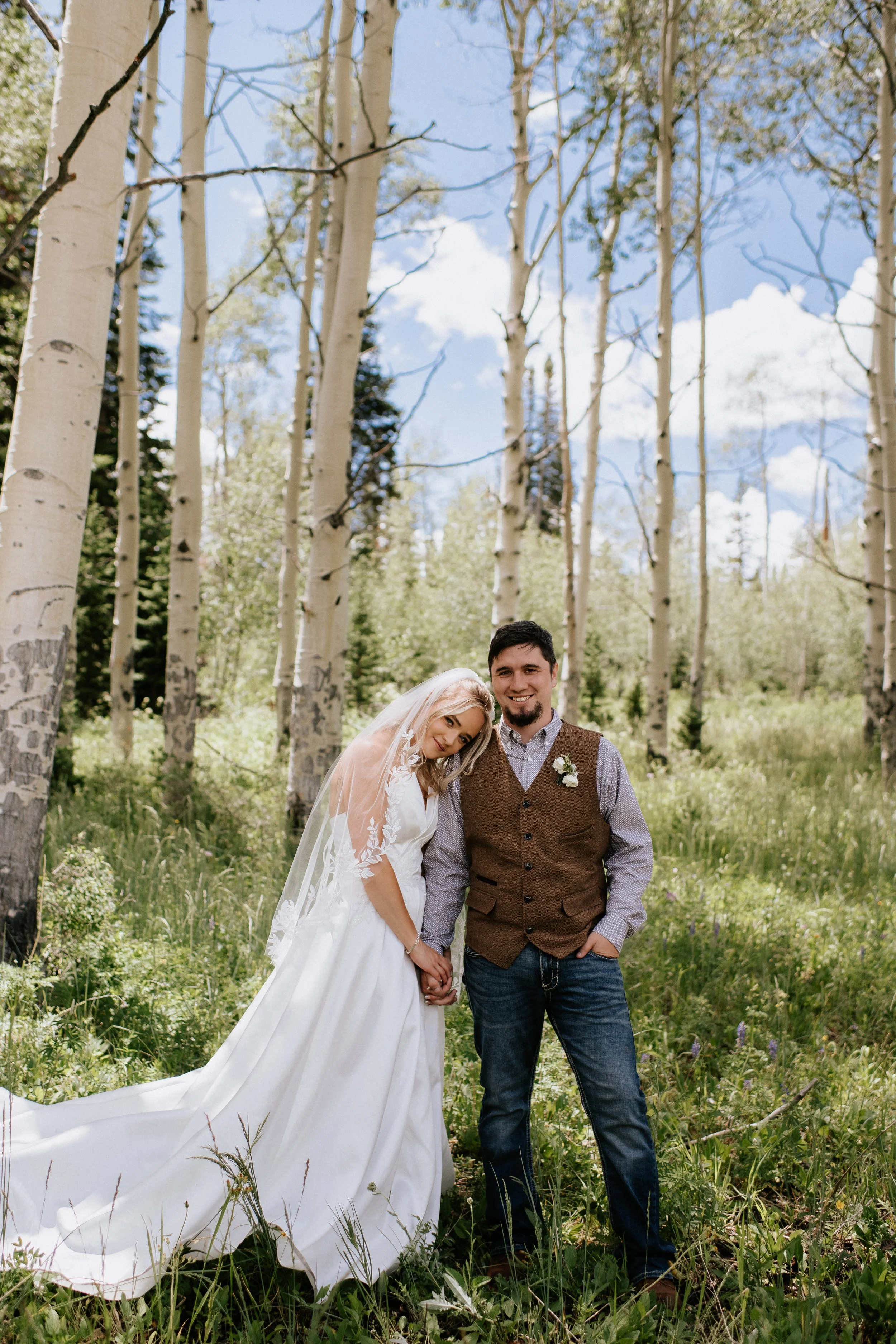 A bride and groom standing in a forest with trees and greenery, smiling and holding hands.