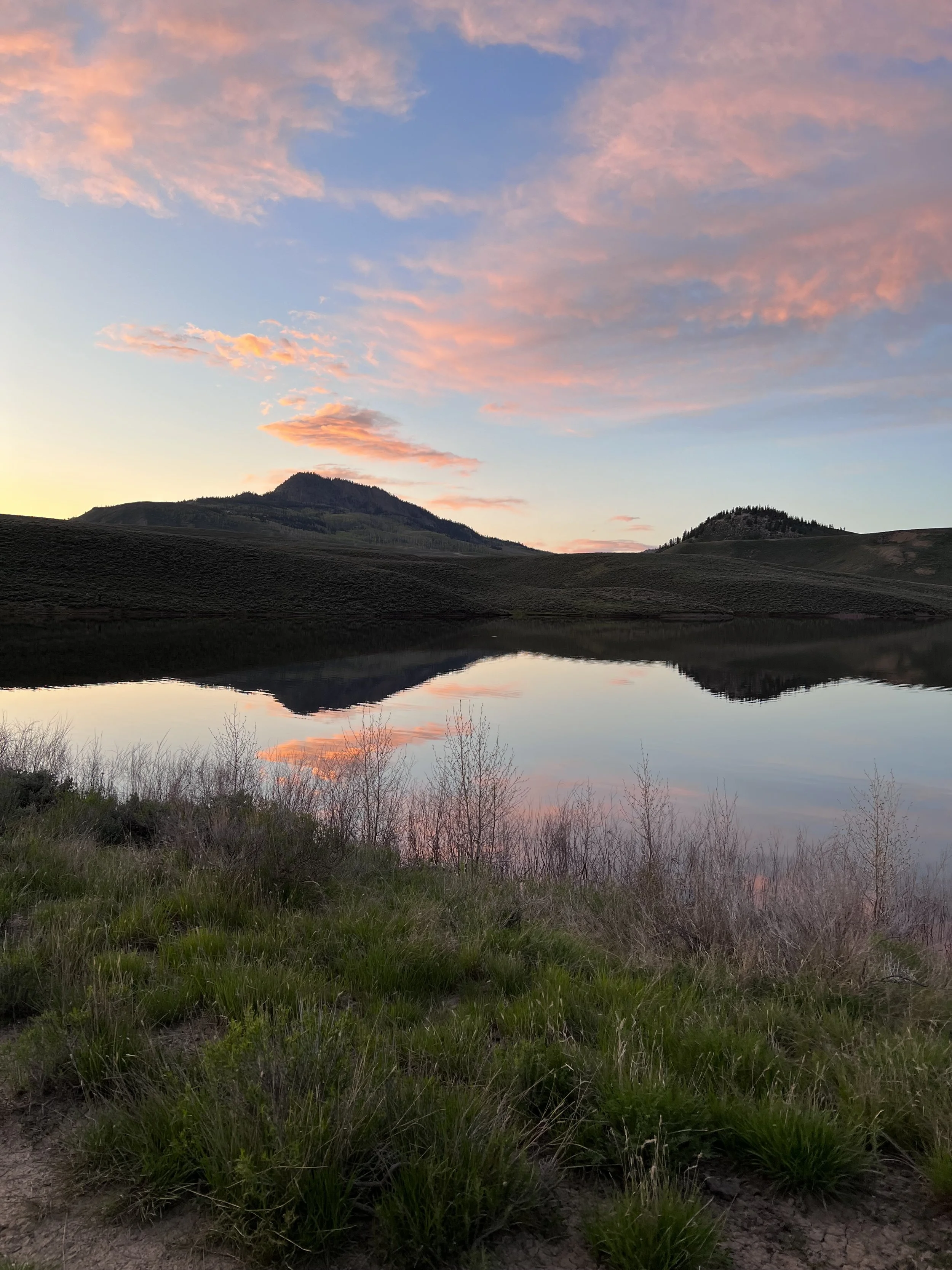 A landscape with a calm lake reflecting the sky, hills, and mountains at sunset or sunrise, with pink and orange clouds and green vegetation in the foreground.
