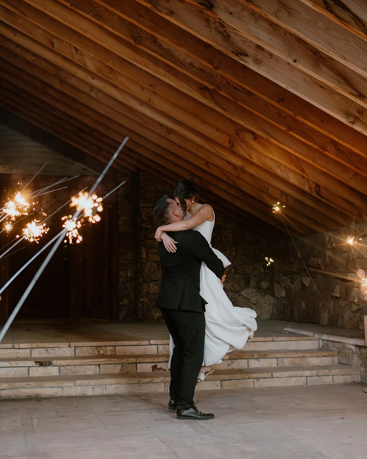A groom lifting and dancing with a bride at a celebration, with sparklers burning around them inside a rustic venue with wooden ceiling and stone walls.