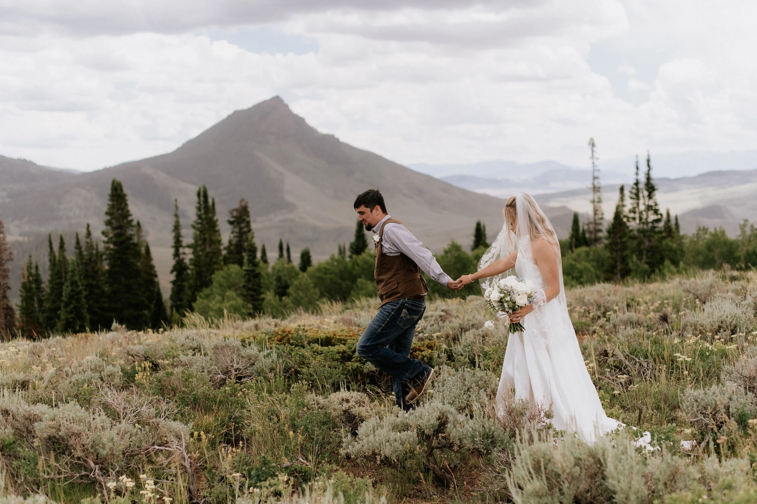 A bride and groom holding hands and walking through a field of low bushes and plants with mountains and trees in the background.