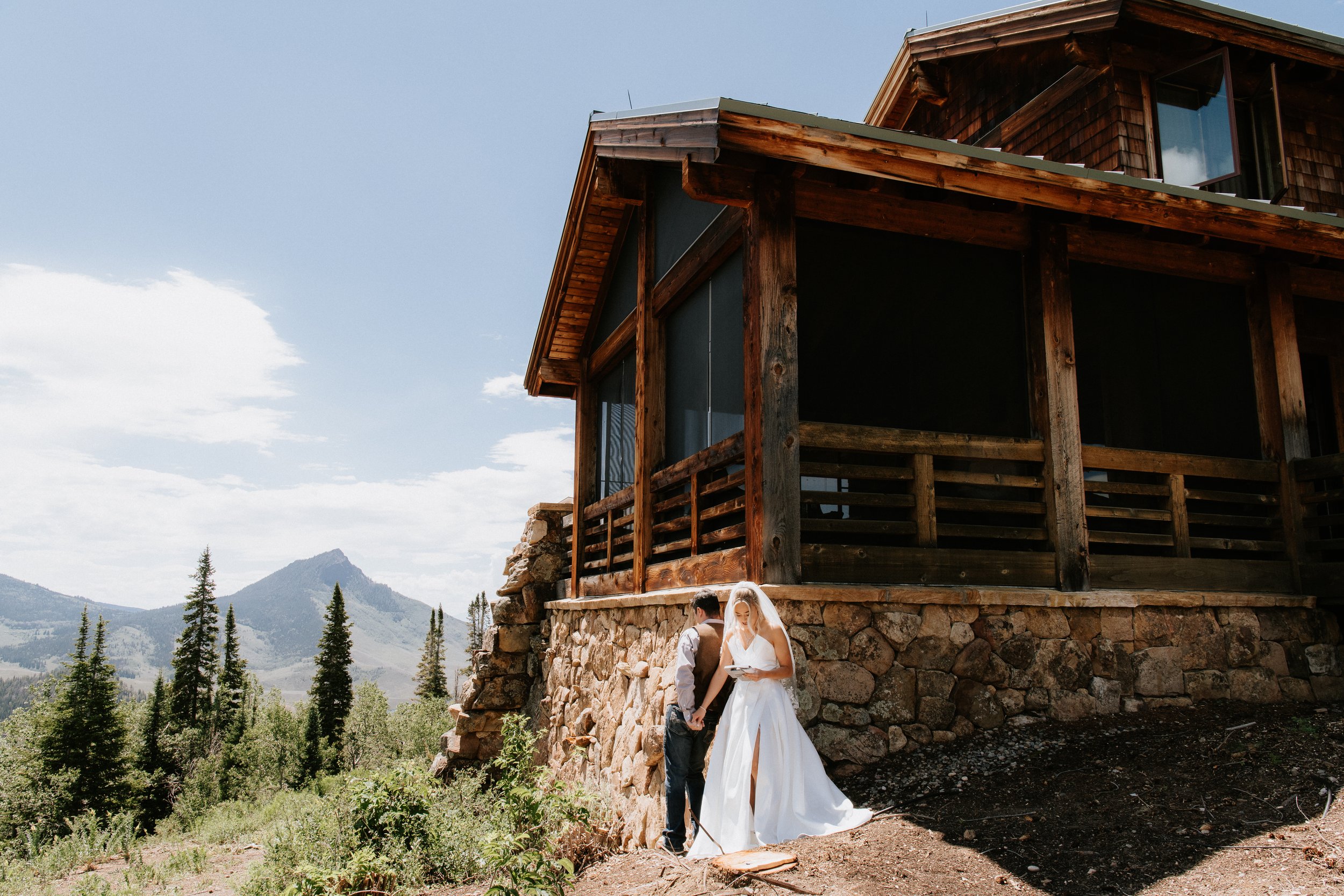 Couple dressed as bride and groom standing outdoors next to a stone foundation with a wooden house on top, in a mountainous forested area during daytime.