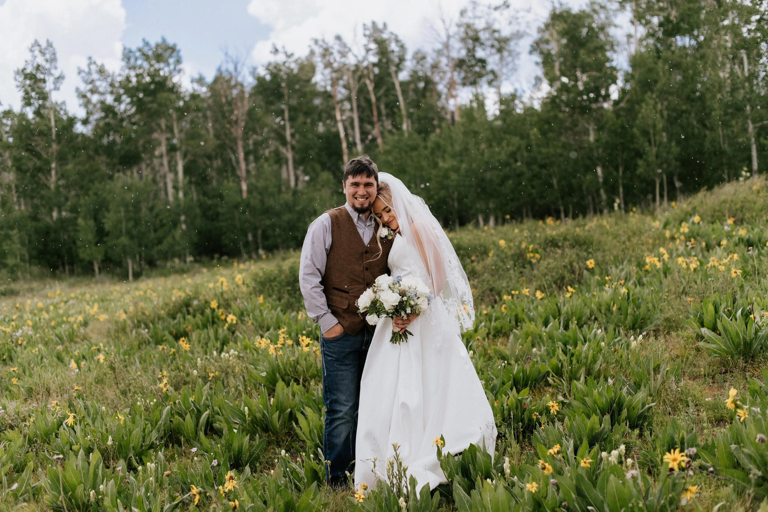 A smiling couple, the bride in a white wedding dress and veil holding a bouquet, and the groom in a brown vest and blue jeans, standing close together in a field of yellow flowers with a background of green trees and a cloudy sky.