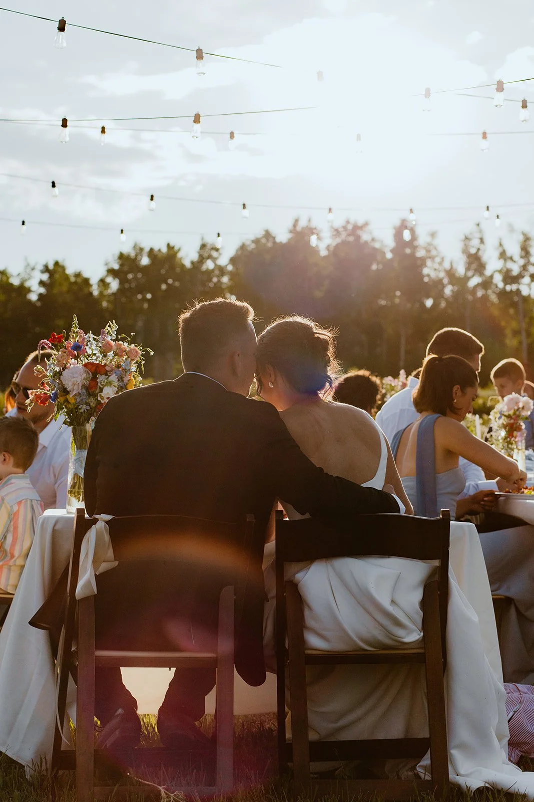 Couple at a wedding reception outdoors during sunset, sitting close together at a table decorated with flowers, with other guests and string lights overhead.