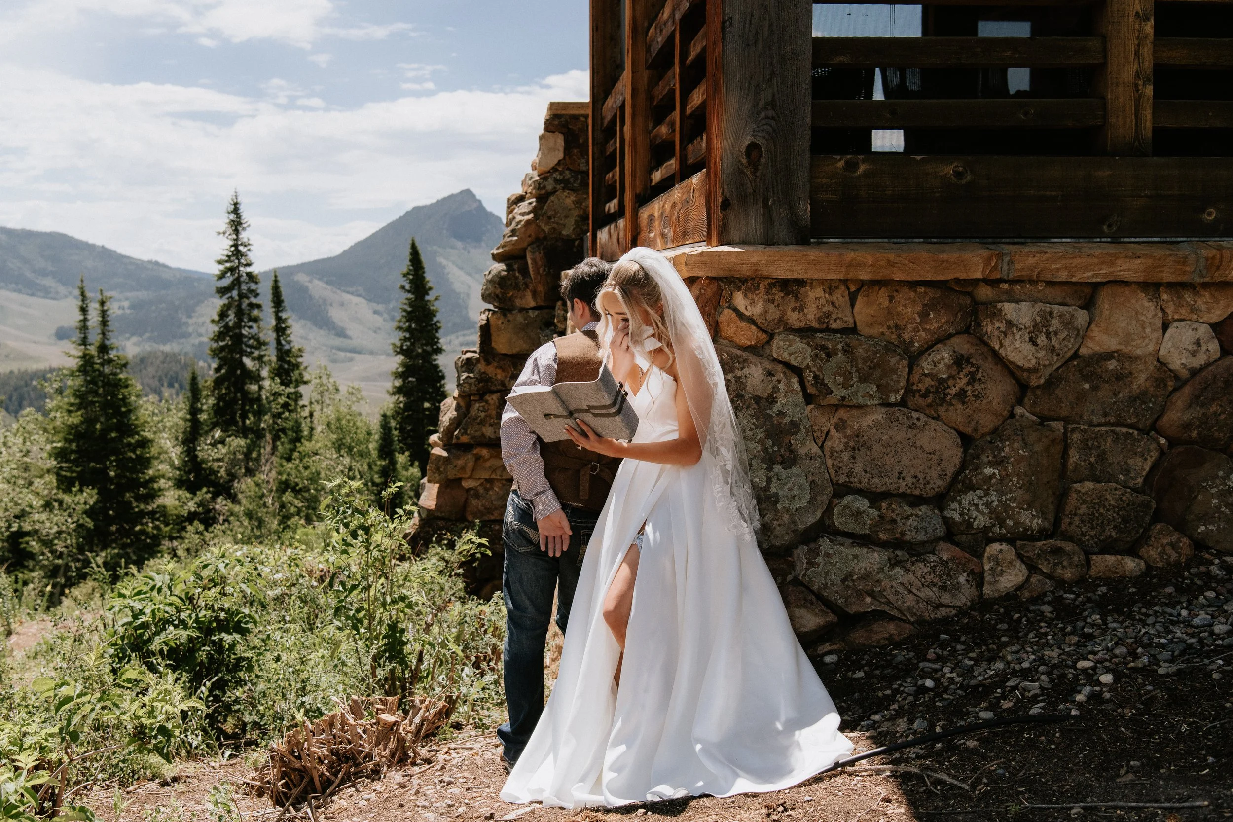 A bride and groom stand outdoors in front of a rustic stone and wood building with mountains and pine trees in the background. The bride wears a white wedding dress with lace details and a veil, while the groom wears a plaid shirt and brown vest. The