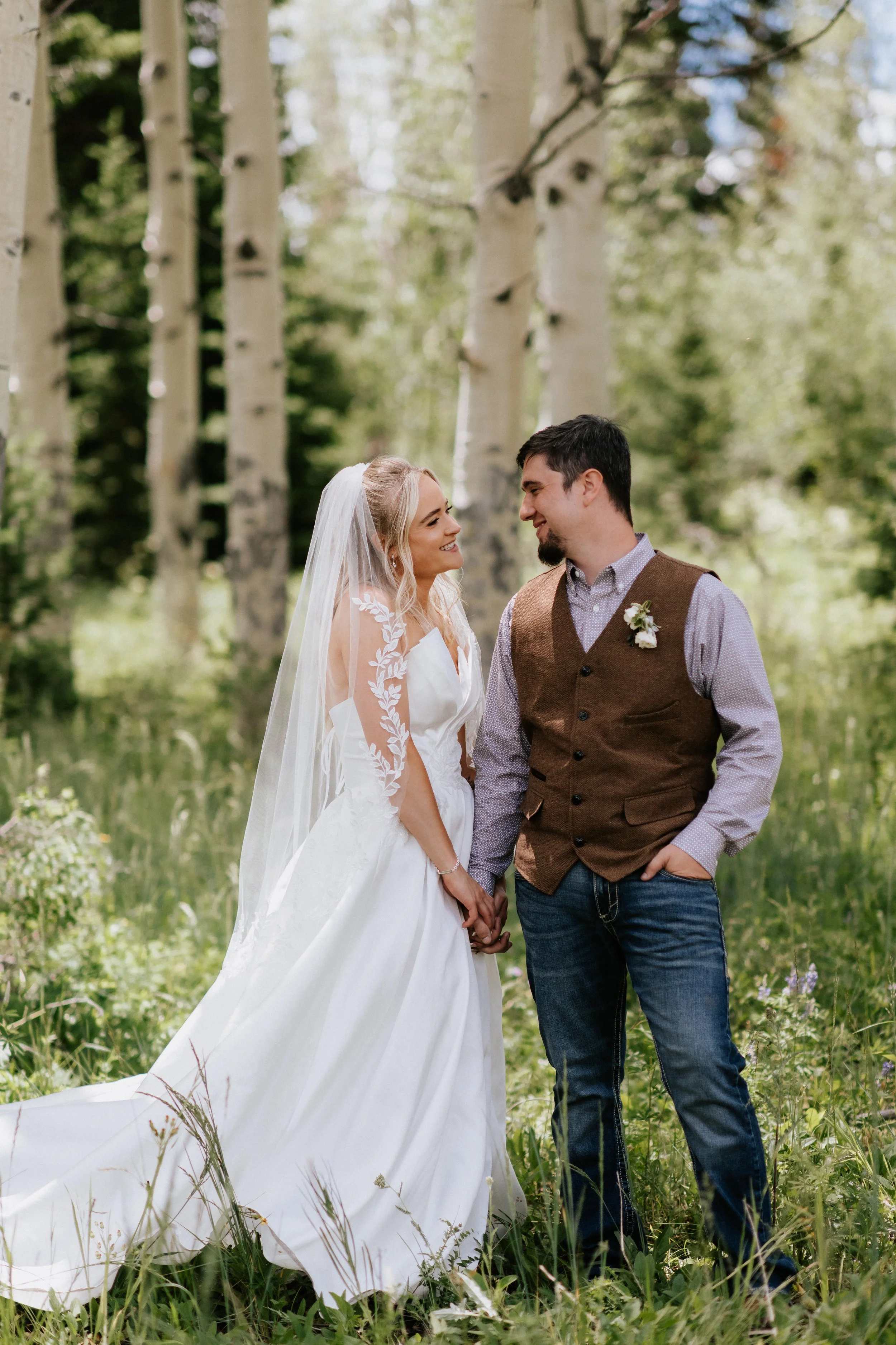 A bride and groom stand hand-in-hand outdoors surrounded by tall trees and greenery, smiling and gazing at each other during their wedding ceremony.