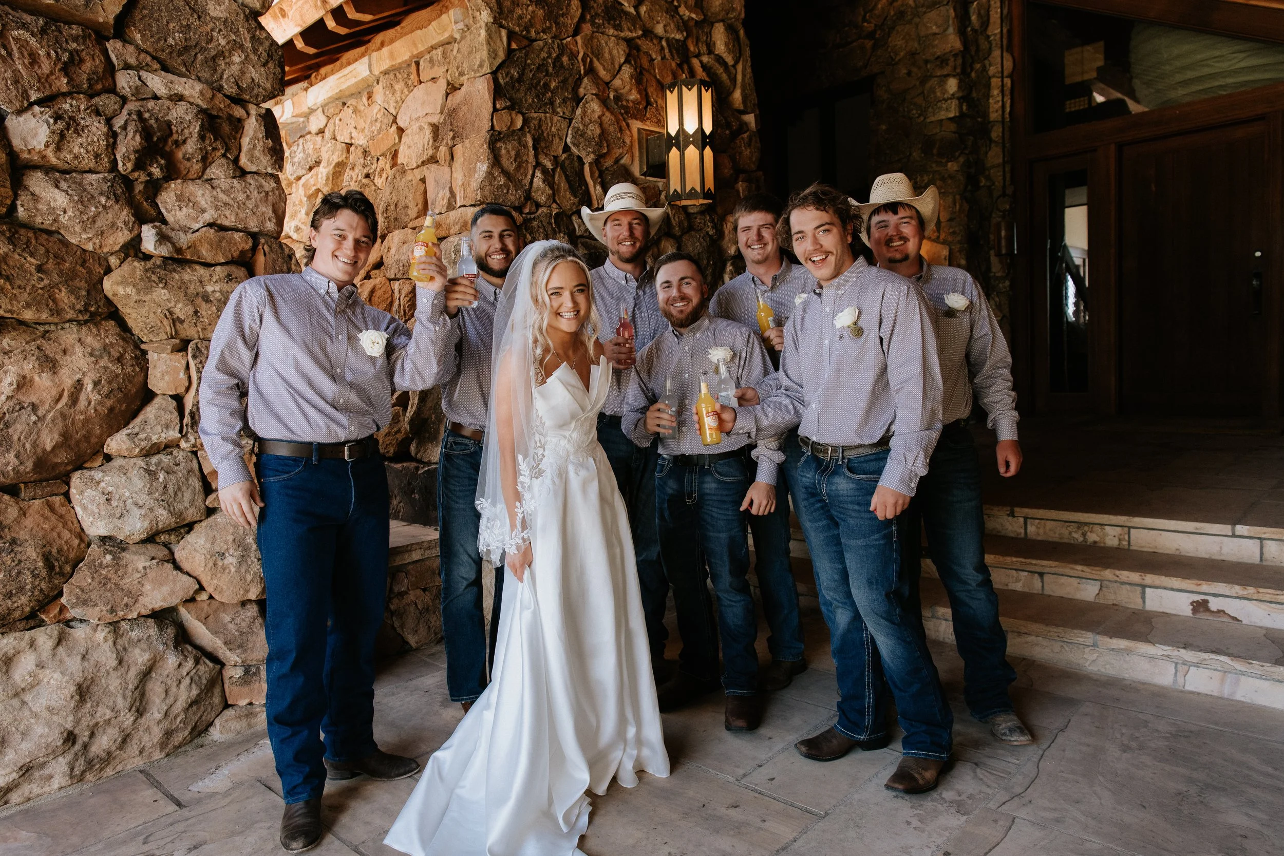A group of ten smiling people, including a bride in a white wedding dress and veil and several men in cowboy hats, standing against a stone wall at a wedding celebration, holding colorful drinks.