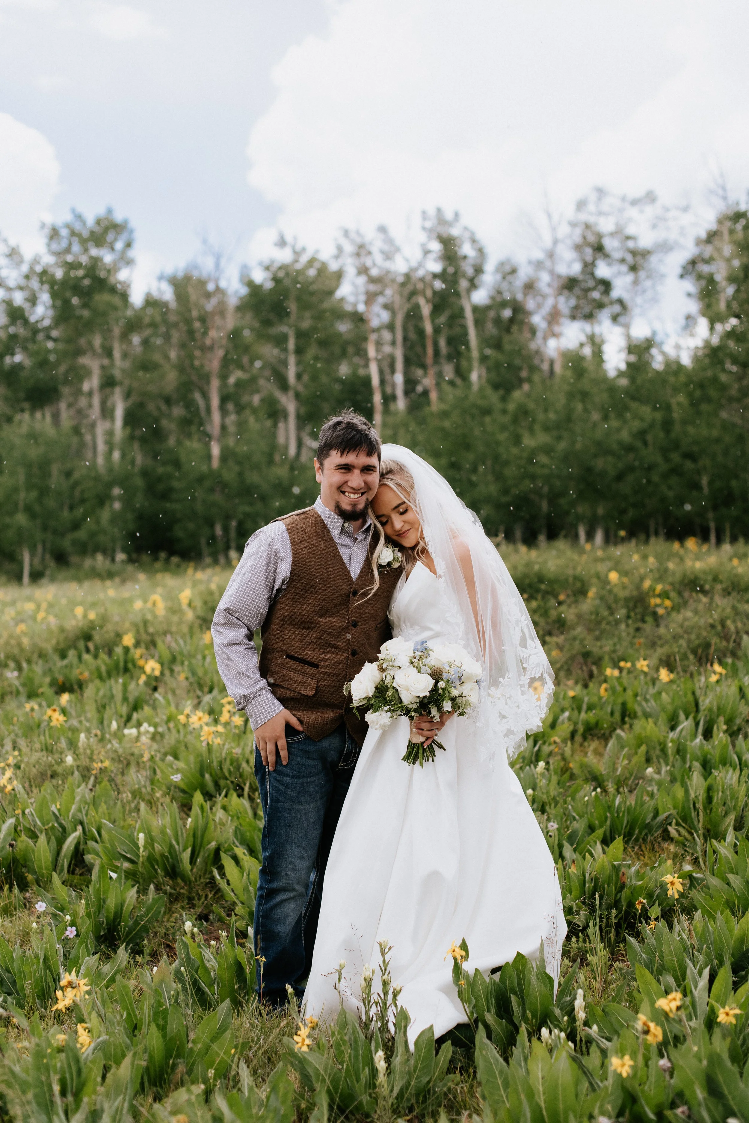 A happy bride and groom pose together in a field of yellow flowers with a forest in the background. The groom wears a brown vest and tie, and the bride holds a bouquet of white flowers, leaning her head on his shoulder.