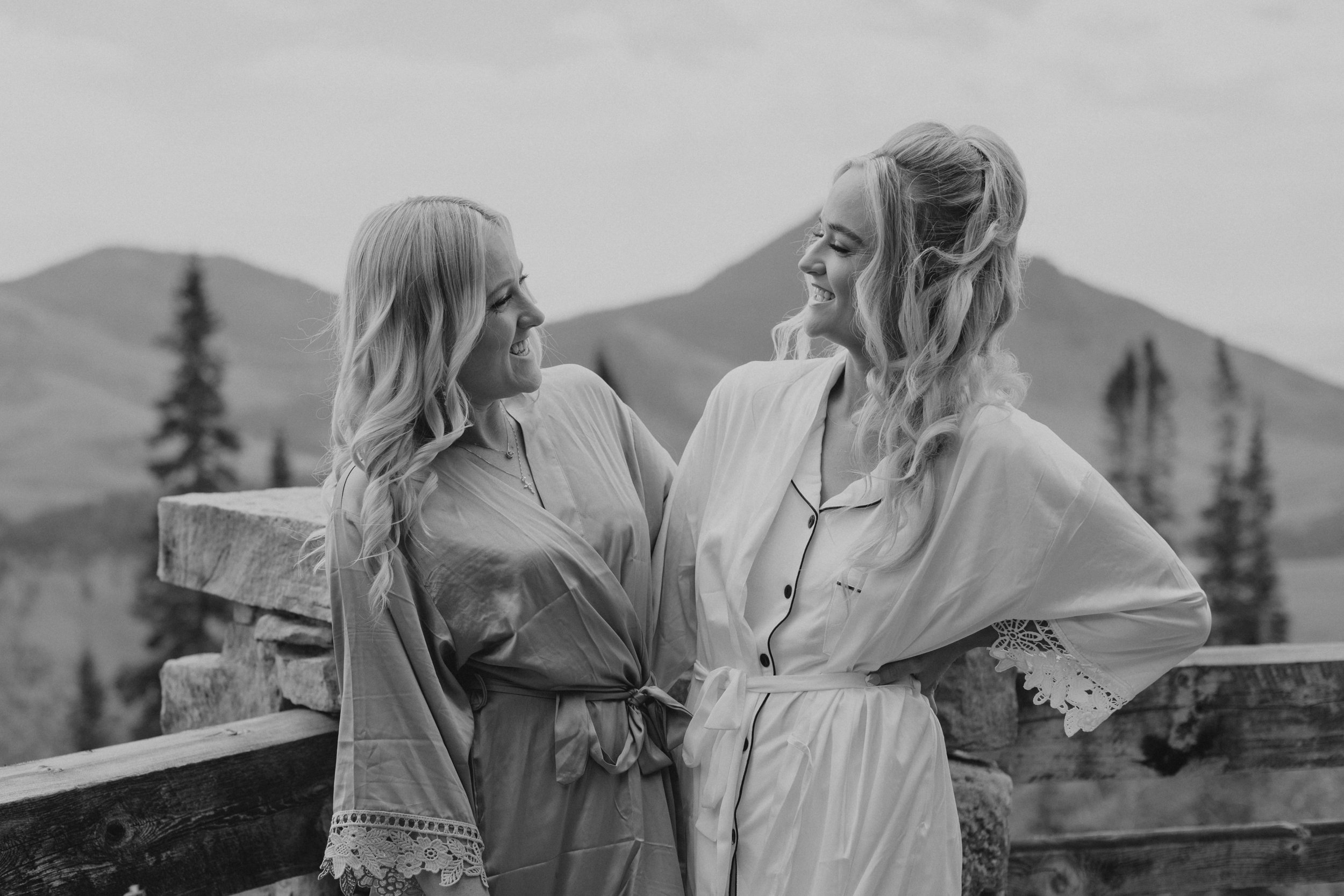 Two women with long, curly blonde hair smiling and looking at each other, standing outdoors in front of mountainous landscape, wearing light-colored clothing.