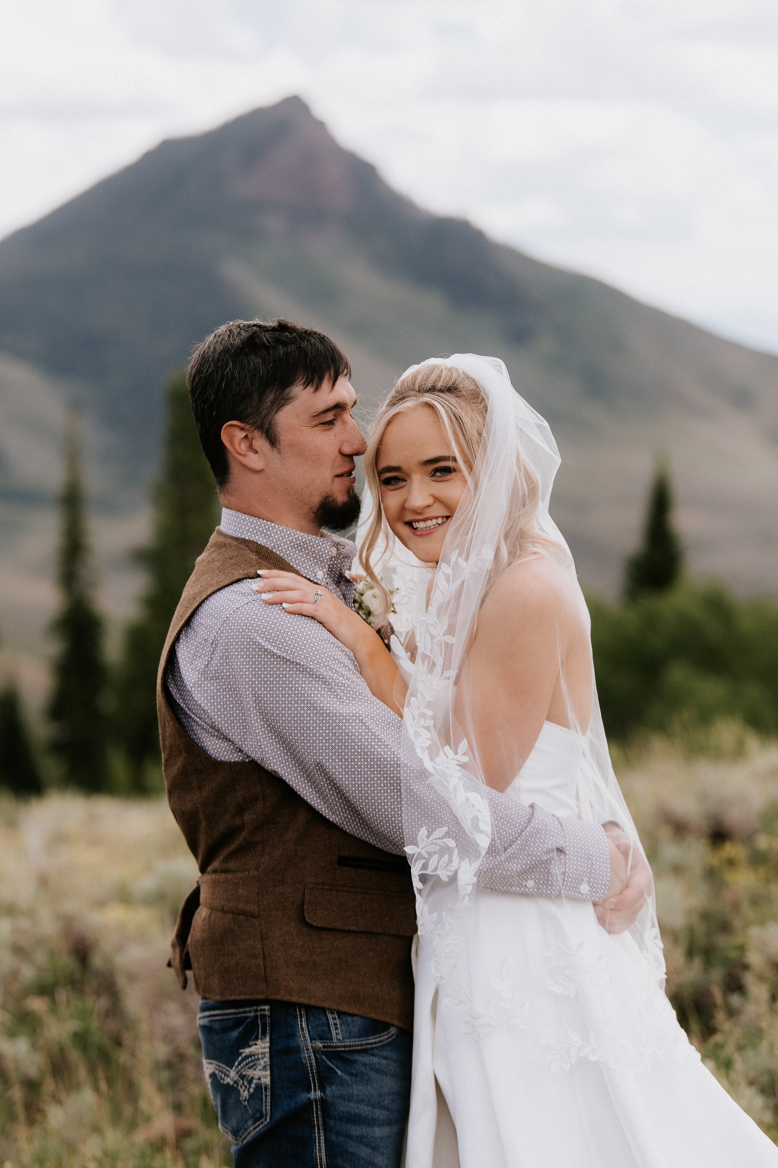 A newlywed couple sharing an embrace outdoors with a mountain in the background. The woman wear a white wedding dress with a veil, and the man is dressed in a brown vest and blue jeans.