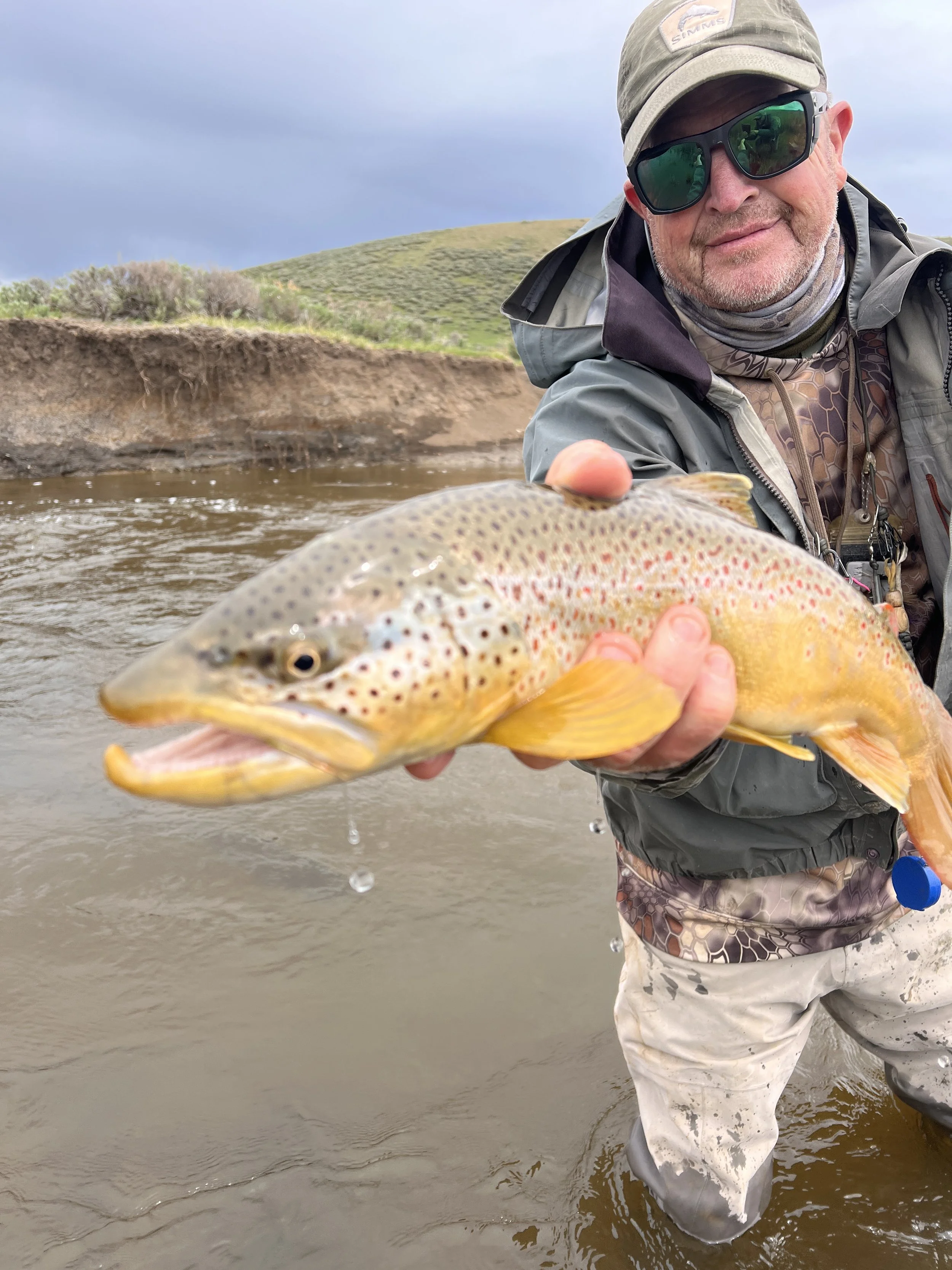 Man in outdoor gear wearing sunglasses, holding a large rainbow trout in a river, with a grassy hill and cloudy sky in the background.