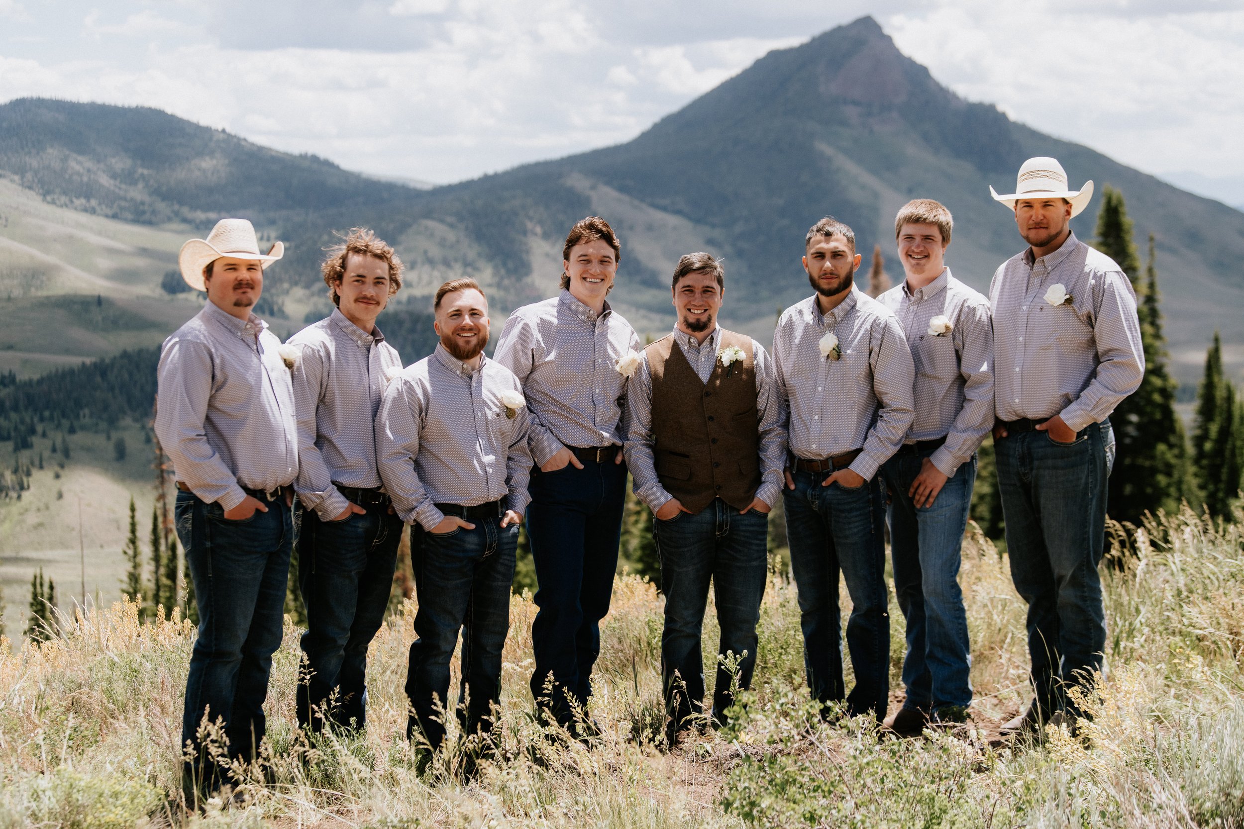 A group of nine young men standing in a field with mountains in the background, dressed in casual button-up shirts and jeans, some wearing white cowboy hats, smiling and posing for a photo.