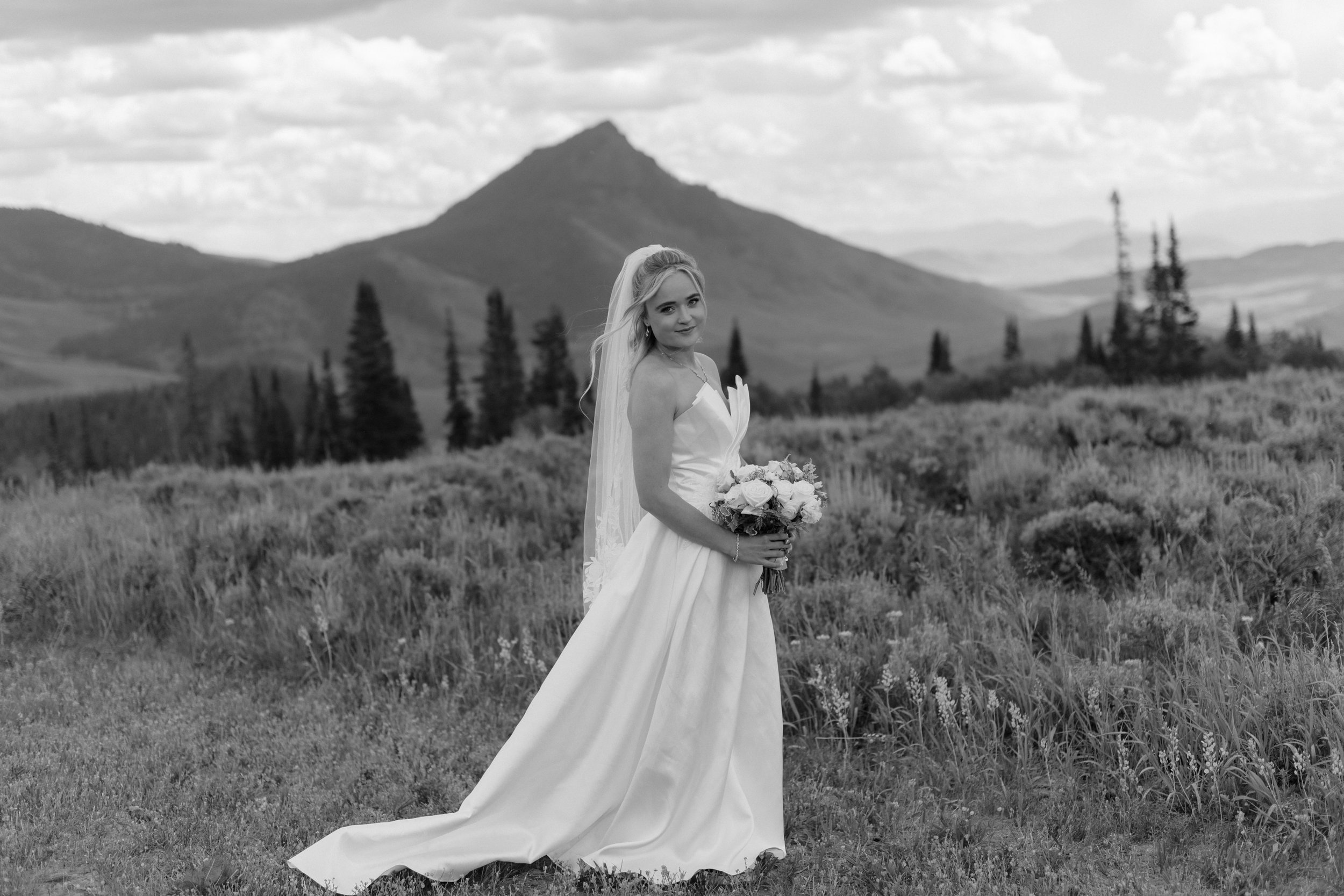 A black and white photo of a woman in a white wedding dress holding a bouquet, standing in a grassy field with trees and mountains in the background.