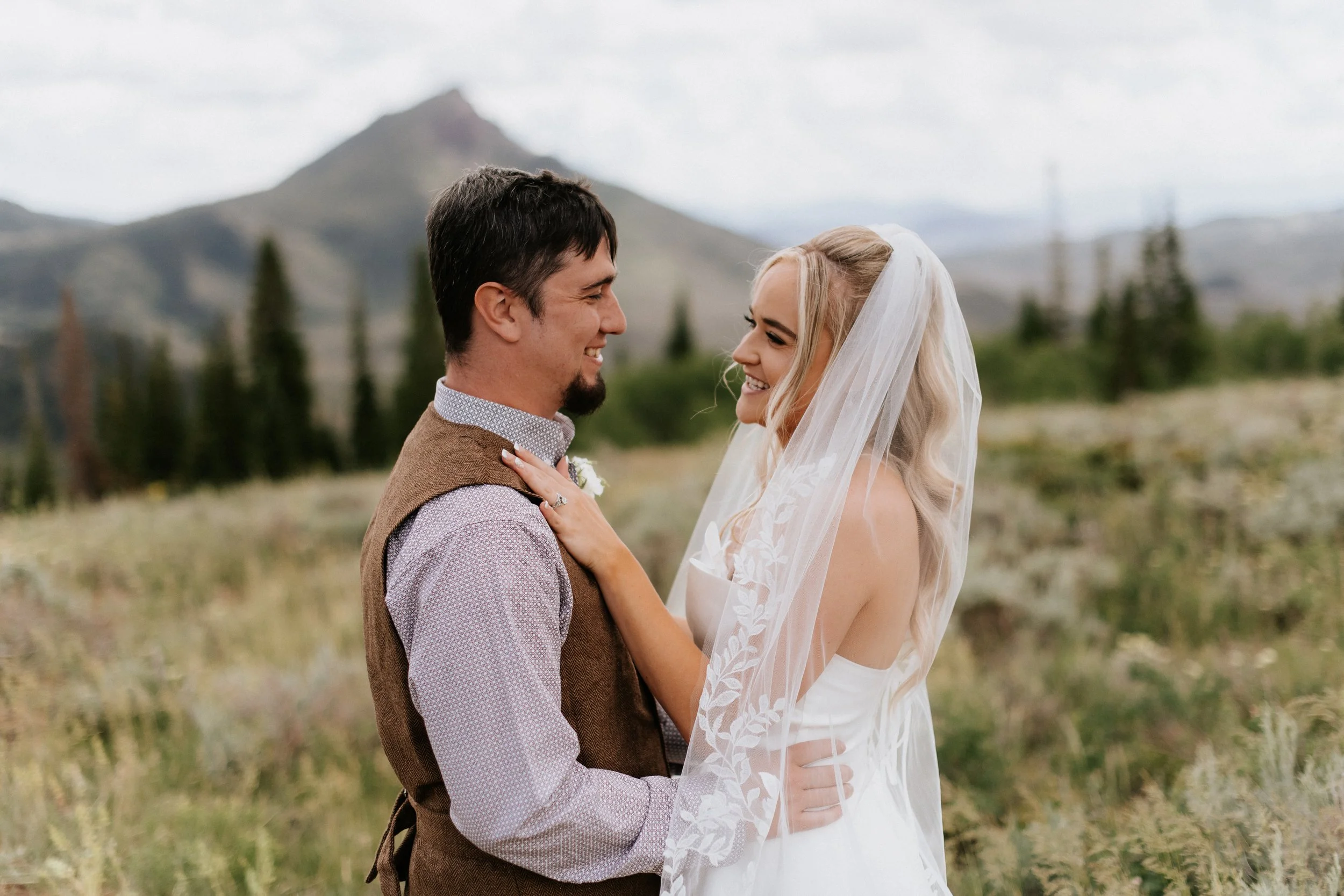 A couple dressed in wedding attire smiling at each other outdoors with mountains and trees in the background.