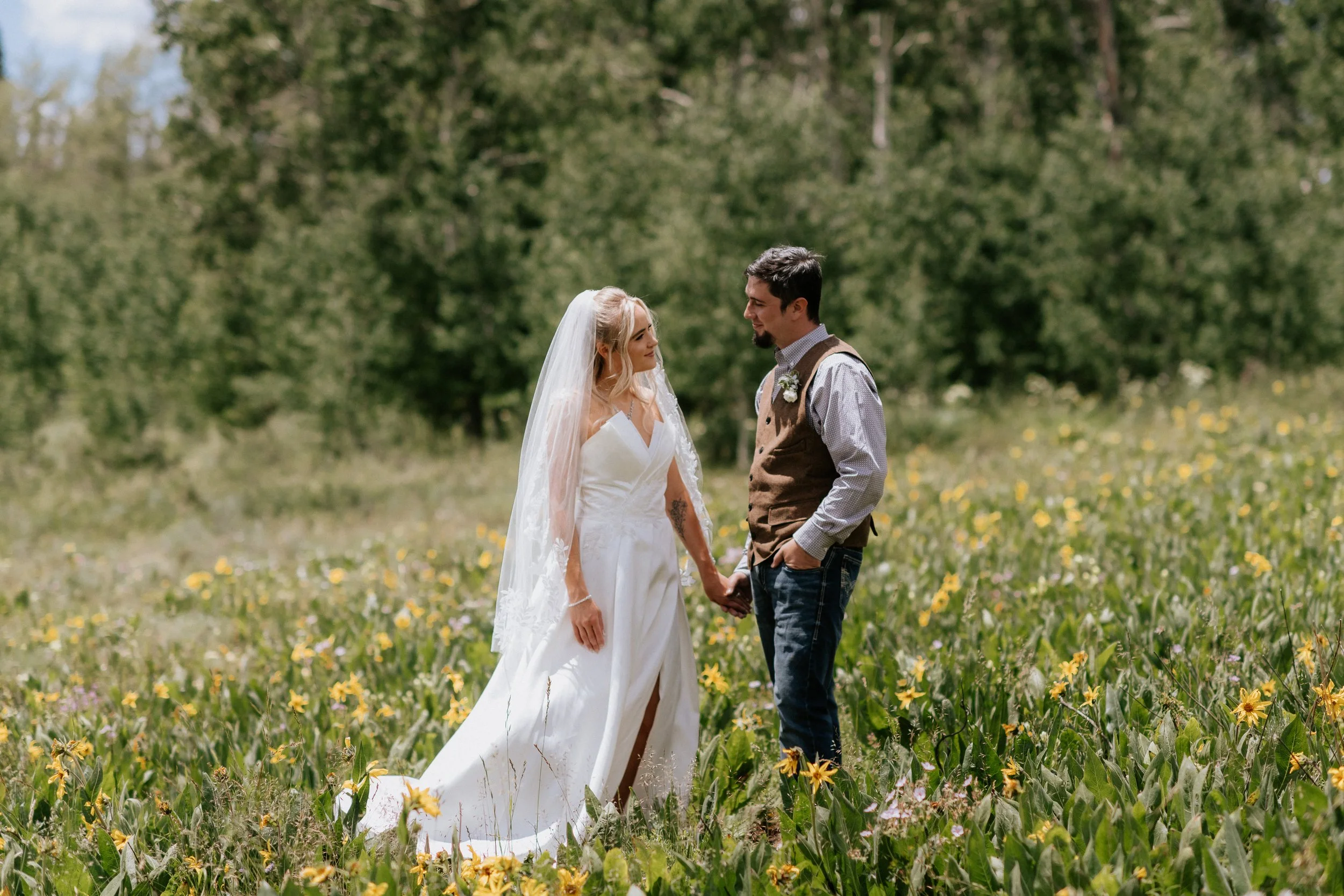 A bride and groom holding hands in a field of yellow flowers with green trees in the background.
