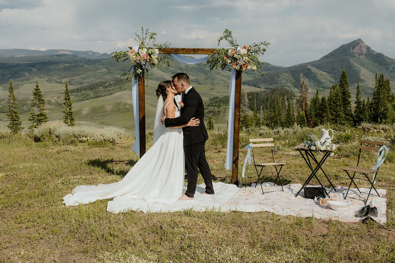 A newlywed couple kisses under a wooden wedding arch decorated with flowers and light blue fabric, outdoors in a scenic mountainous landscape.