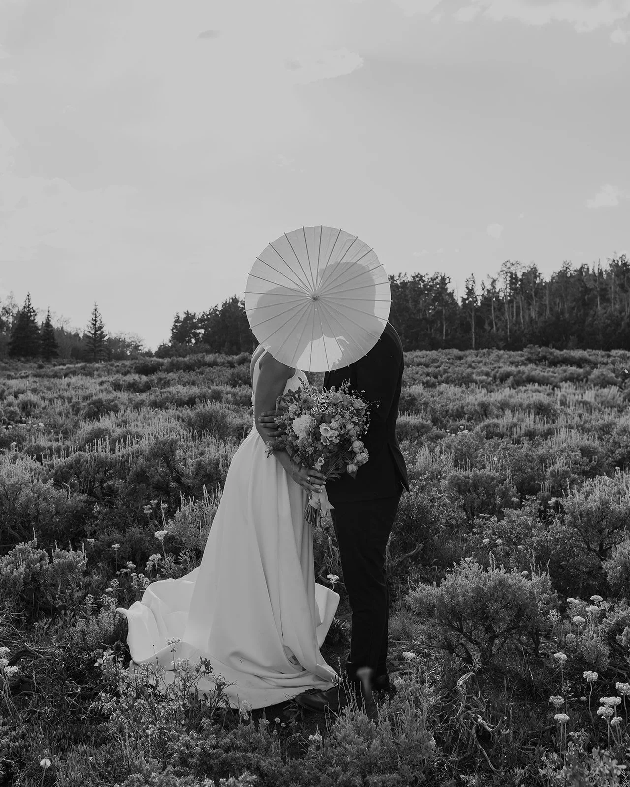 A bride and groom standing close together in a field, with the bride holding a bouquet of flowers. The couple is kissing with an umbrella covering their faces. The photo is in black and white.