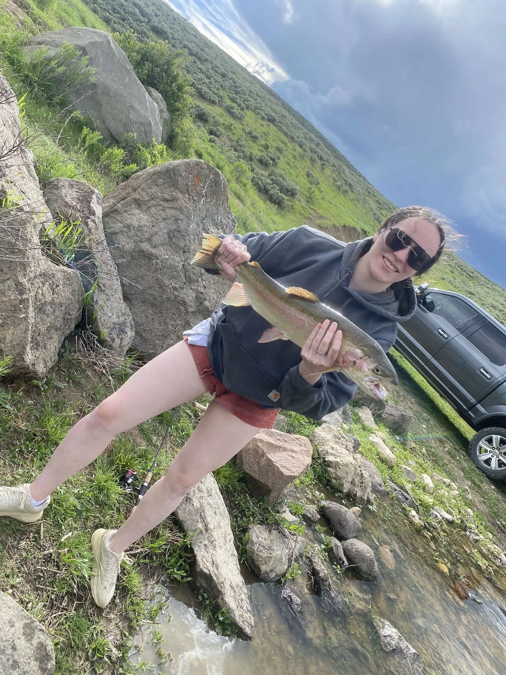 Woman in sunglasses holding a large fish, standing near rocks on a riverbank with a grassy hill and a parked black truck in the background.