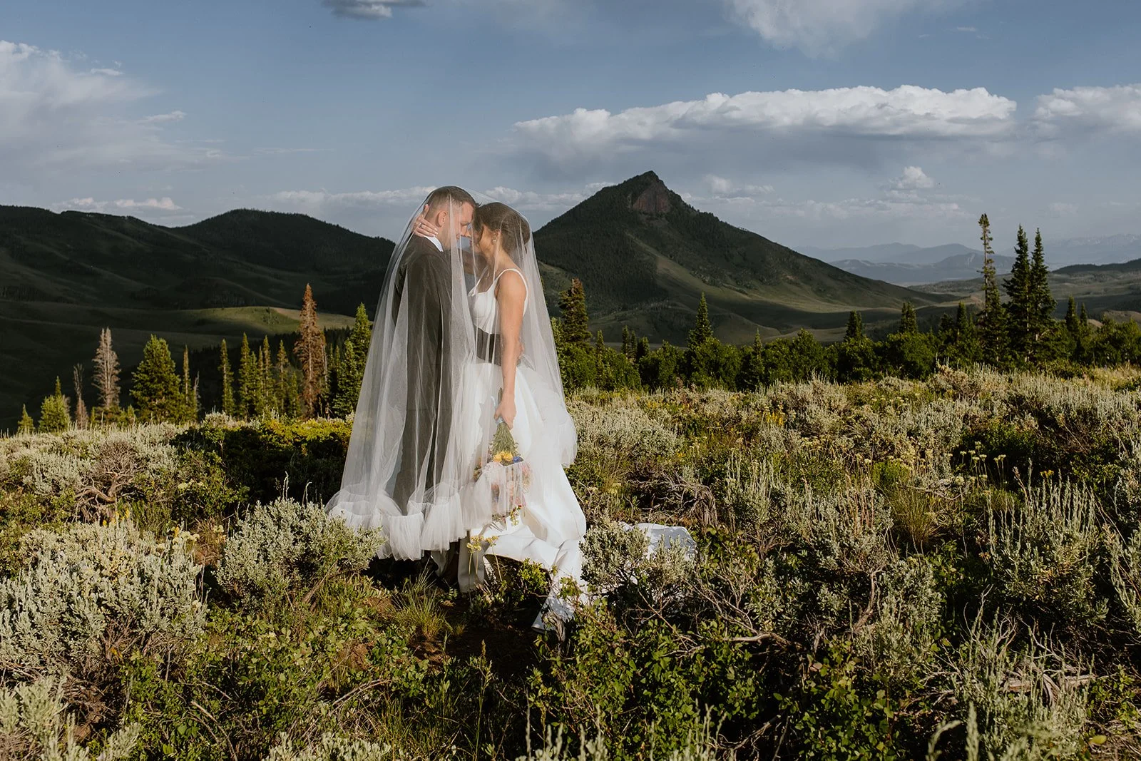 A bride and groom dressed in wedding attire share an intimate moment in a scenic outdoor landscape with mountains, trees, and clouds in the background.