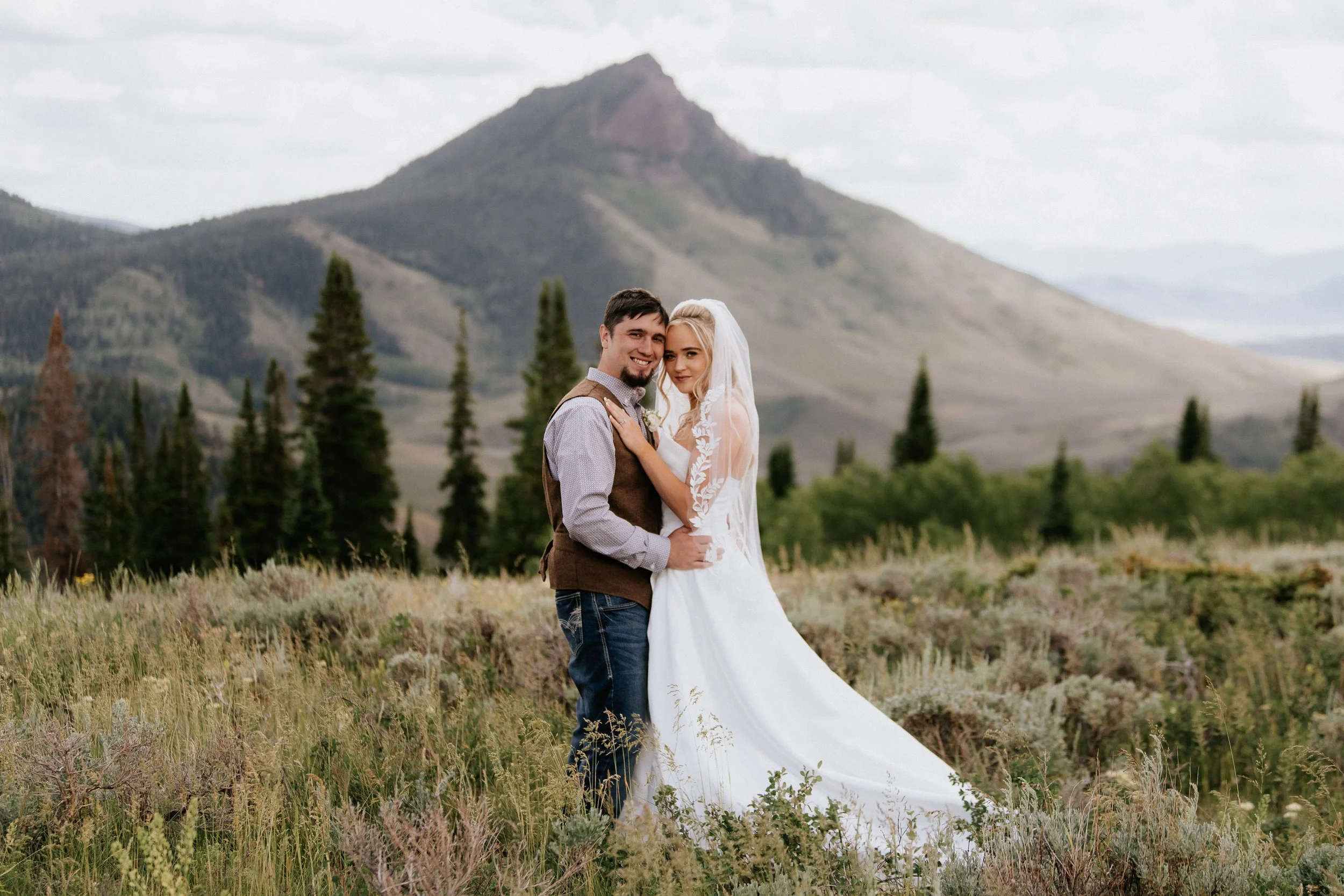 A bride and groom standing together in a grassy field with a mountain and tall pine trees in the background.