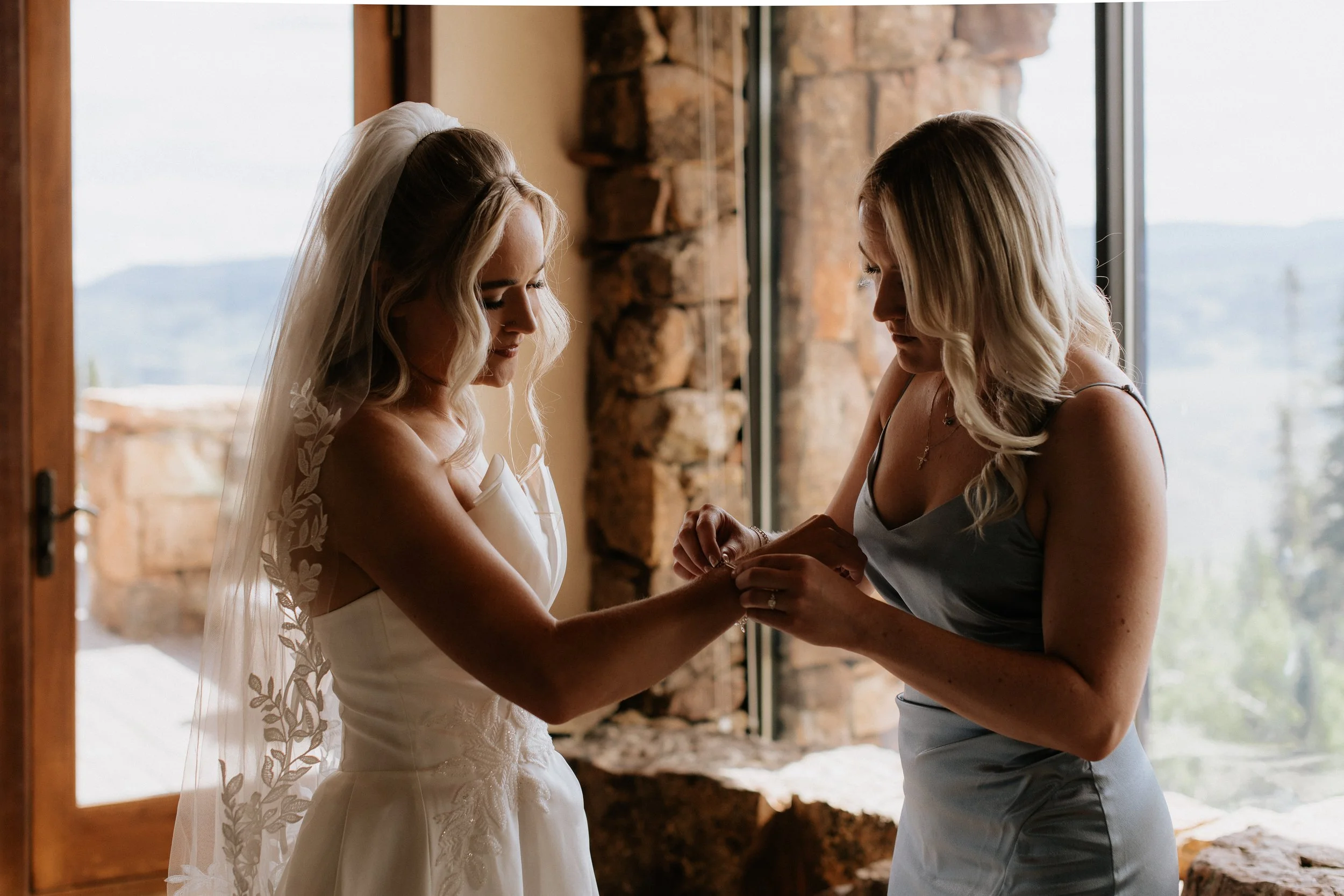 A bride in a white wedding dress with lace details and a veil, standing inside by a large window, while another woman, possibly her bridesmaid, helps her put on a ring or bracelet, with a rustic stone wall and view of outdoors in the background.