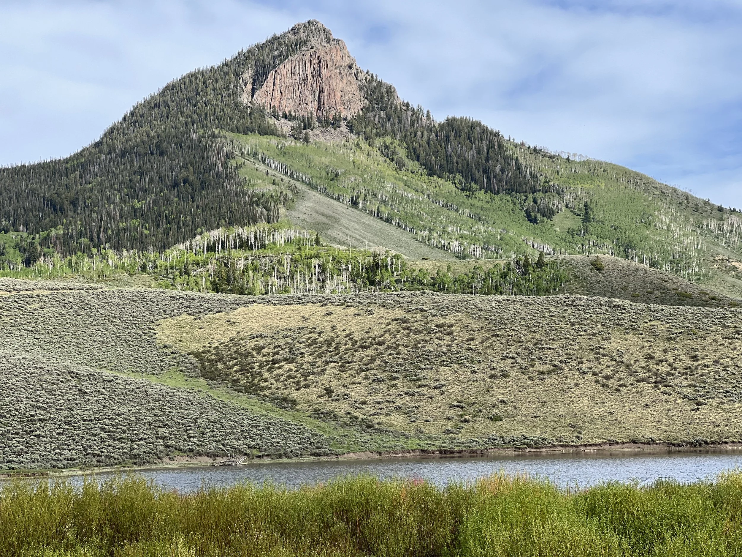 Scenic view of a mountain with lush green forests and a rocky peak, a grassy hillside, a small body of water at the base, and a partly cloudy sky.
