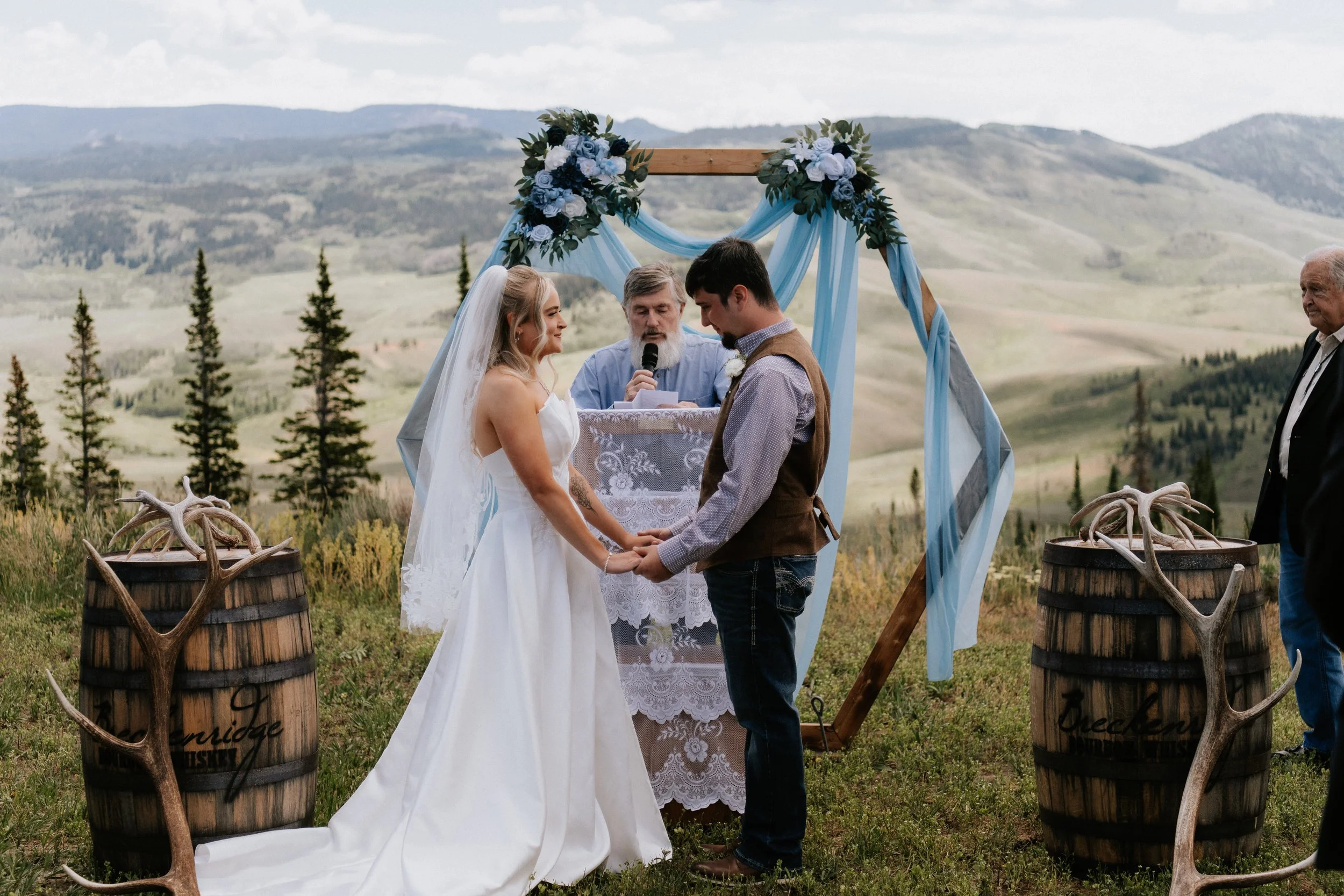A couple getting married outdoors, holding hands and facing each other with an officiant standing behind a decorated altar, in a scenic mountainous landscape.