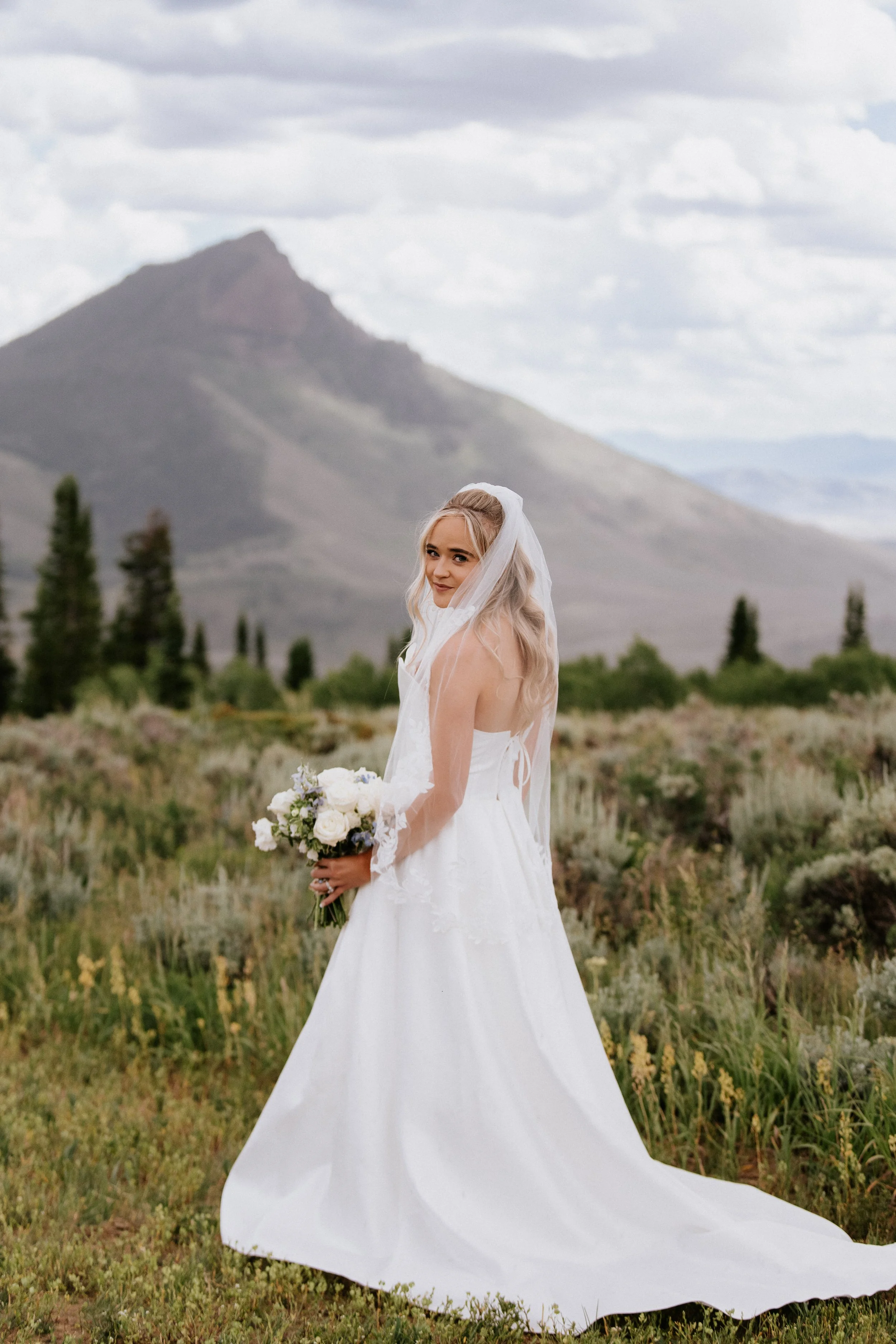 A bride in a white wedding dress with lace details, holding a bouquet of white flowers, stands outdoors with a mountain and cloudy sky in the background.