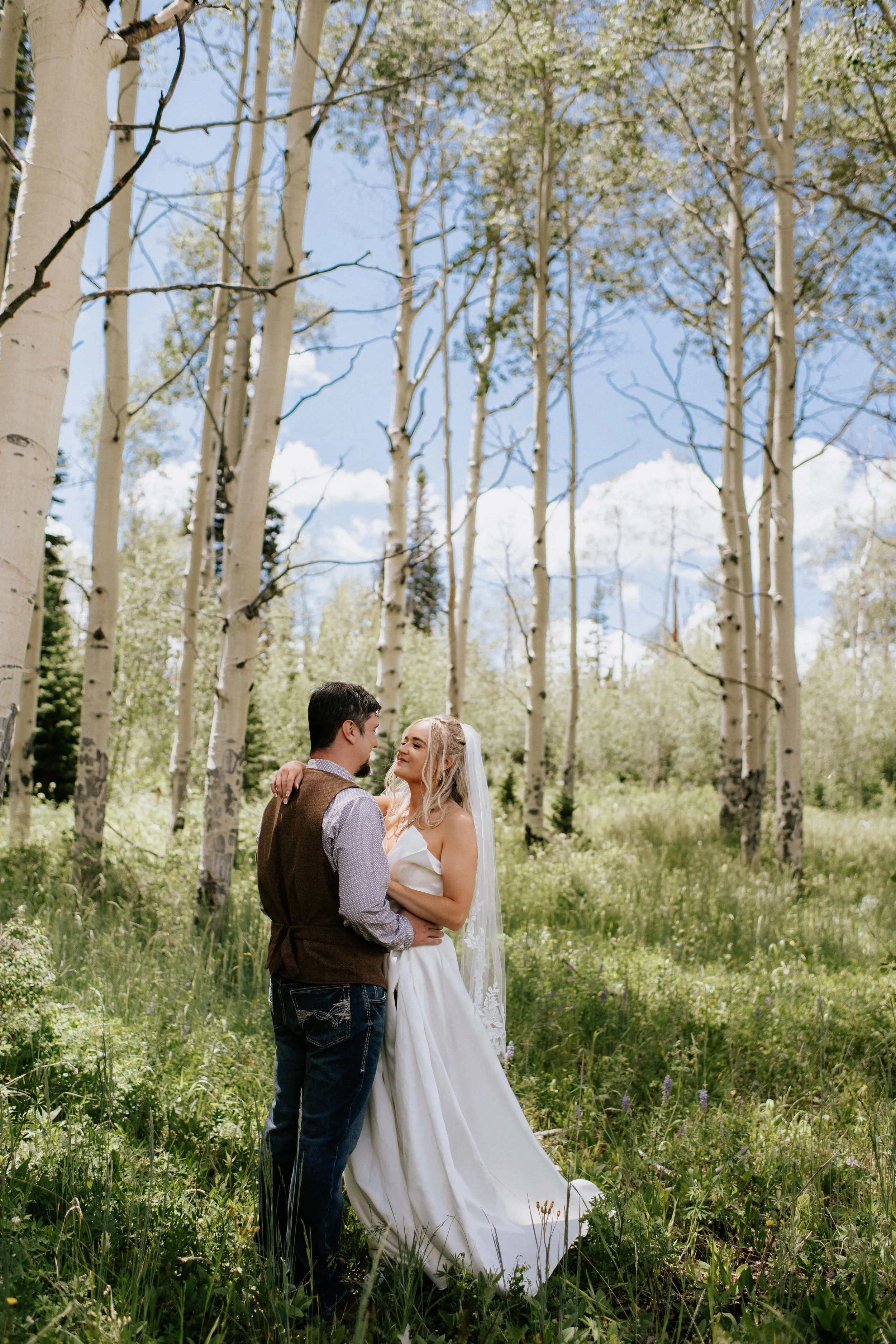 A bride and groom embrace in a forest clearing with tall aspen trees and green grass, during a sunny day.