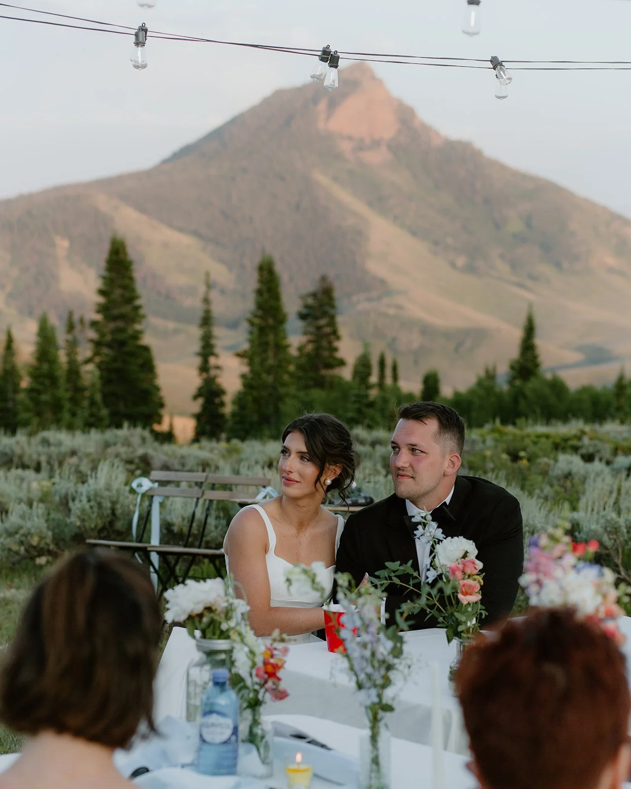 A bride and groom sitting at a wedding reception table outdoors with mountains and pine trees in the background, decorated with flowers and candles.