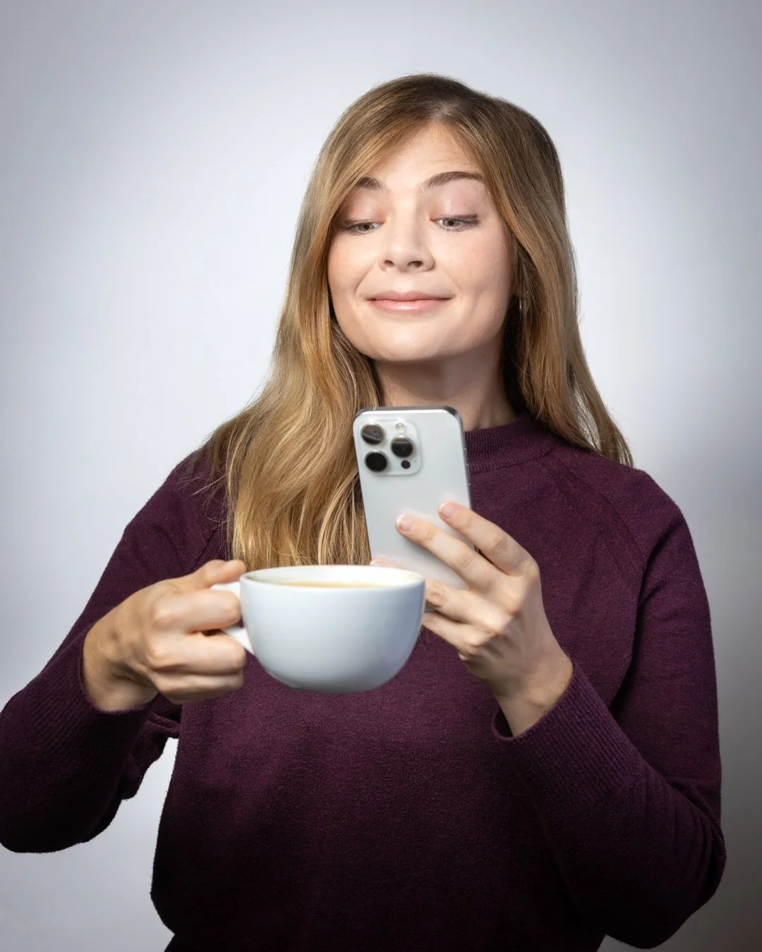 A woman with long red hair holding a white mug of coffee and looking at her smartphone.