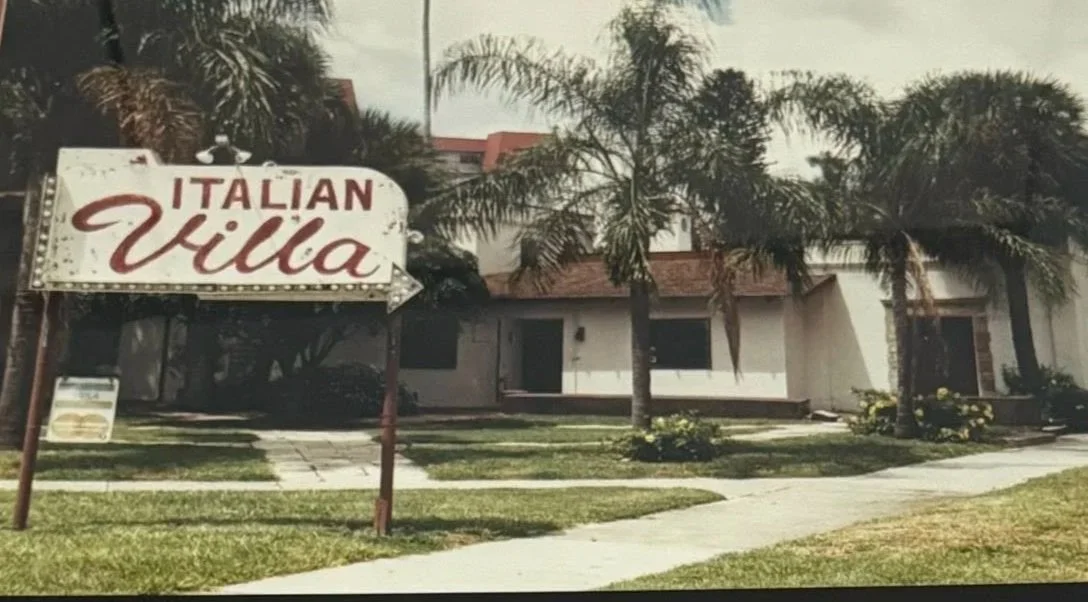A sign reading 'Italian Villa' points toward a building with white walls, a red-tiled roof, and a lawn with palm trees.