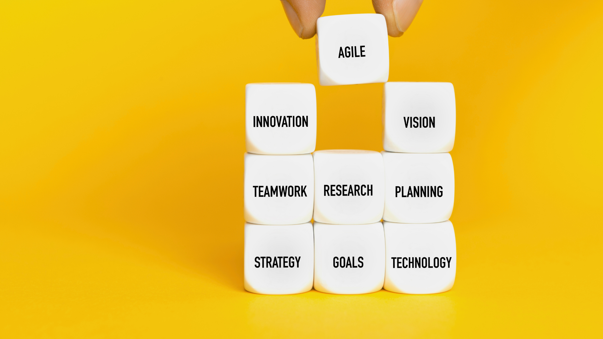 A hand arranging white cubes with black text on a yellow background. The cubes display words like 'AGILE', 'INNOVATION', 'VISION', 'TEAMWORK', 'RESEARCH', 'PLANNING', 'STRATEGY', 'GOALS', and 'TECHNOLOGY', representing concepts related to business and project management.