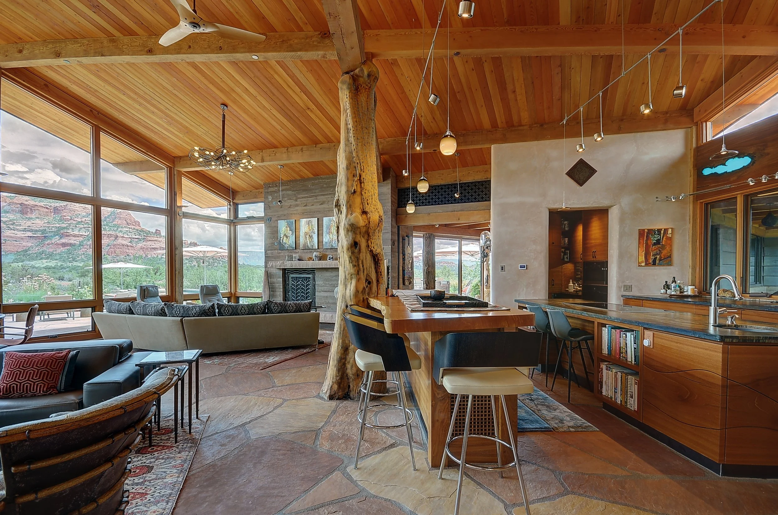 Open-concept living space with large windows, wood ceiling, stone flooring, and modern furniture including a sofa, chairs, and a kitchen island with bar stools. A large driftwood log acts as a central support, surrounded by scenic mountain view.