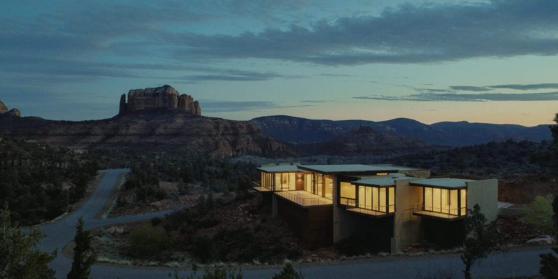 Modern house with large windows and balconies situated on a rugged landscape with trees, mountains, and a sunset sky in the background.