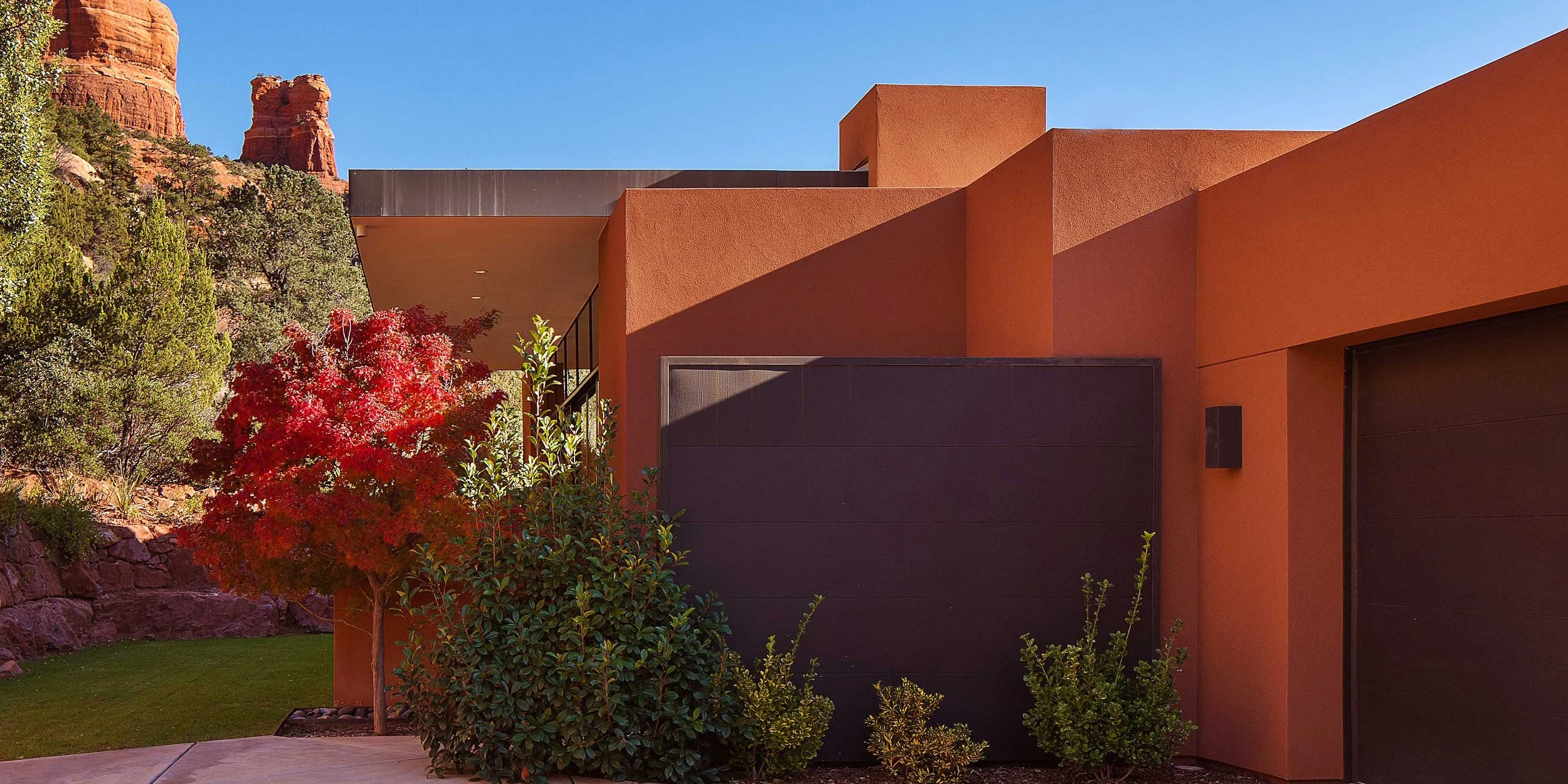 Modern house with orange stucco walls, black garage doors, and surrounding desert landscaping including a red-leafed tree, green bushes, and some rocks, with red rock formations in the background under a clear blue sky.