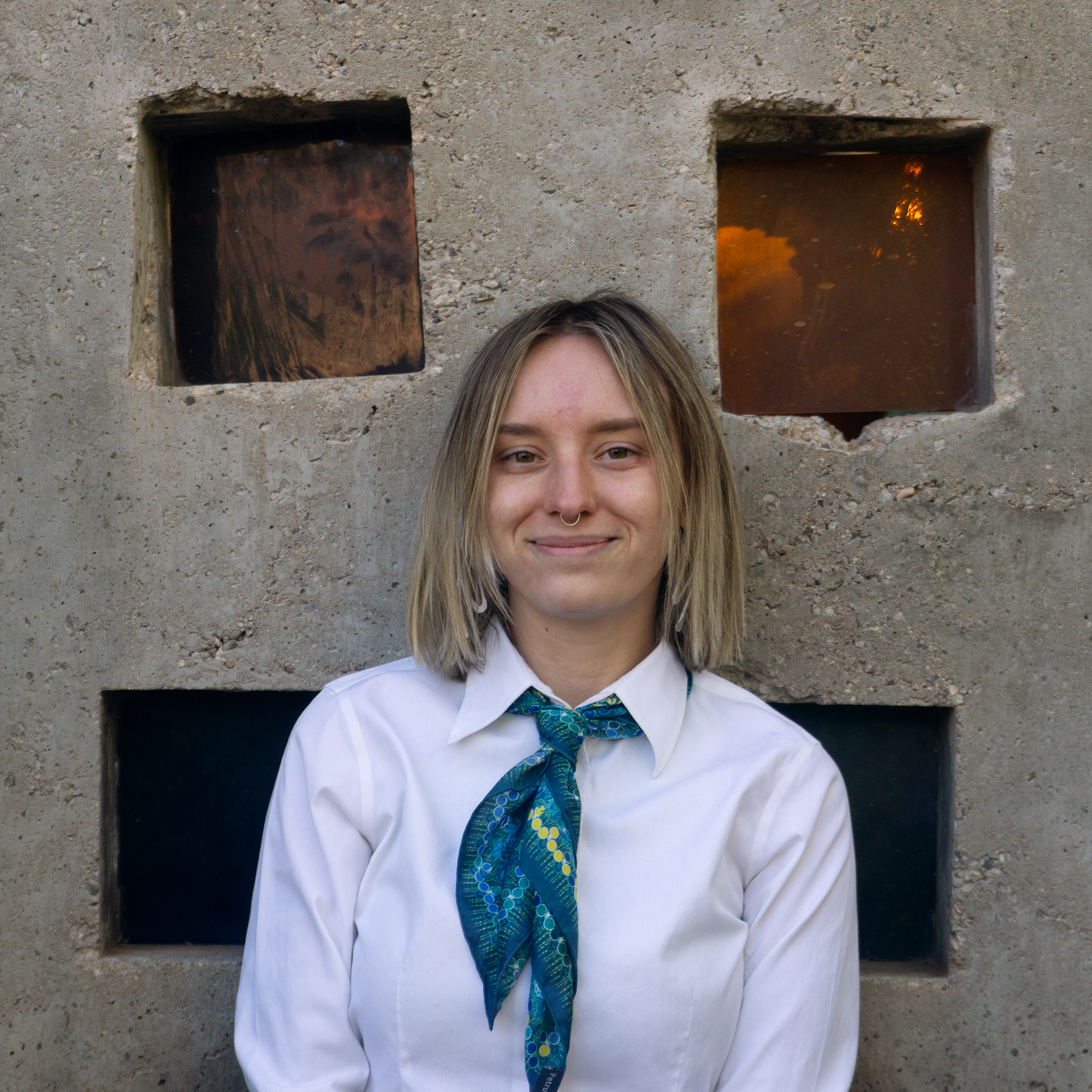 A woman with shoulder-length blonde hair, wearing a white shirt and a blue patterned scarf, standing in front of a textured concrete wall with square windows.