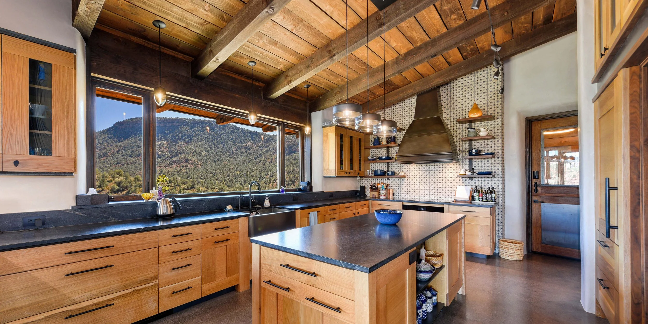 Rustic kitchen with wooden cabinets, black countertops, large window showing mountains, and exposed wooden beams on ceiling.