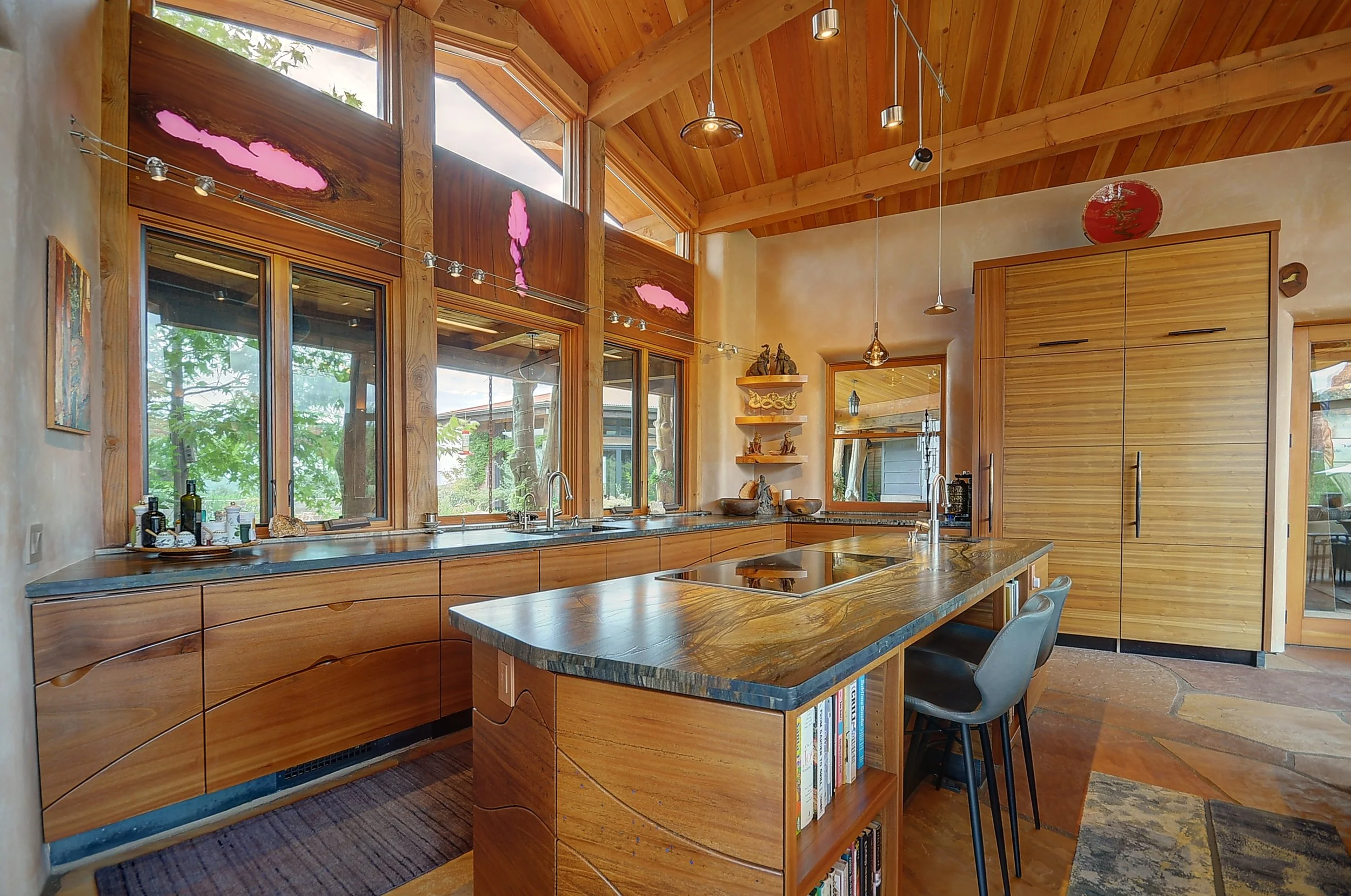 A kitchen with custom cabinetry and large windows framed by timber with pink inlay, a marble countertop, open shelves with decorative items, and a small bookshelf on the kitchen island.