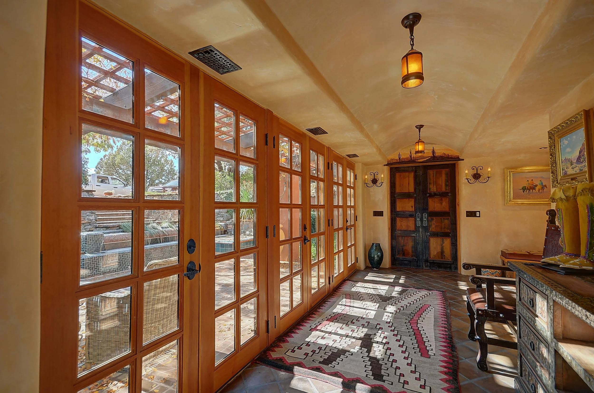 Entryway with large wooden framed glass double doors, a colorful patterned rug, and a small side table with artwork and decorative pillows, illuminated by warm ceiling lights.