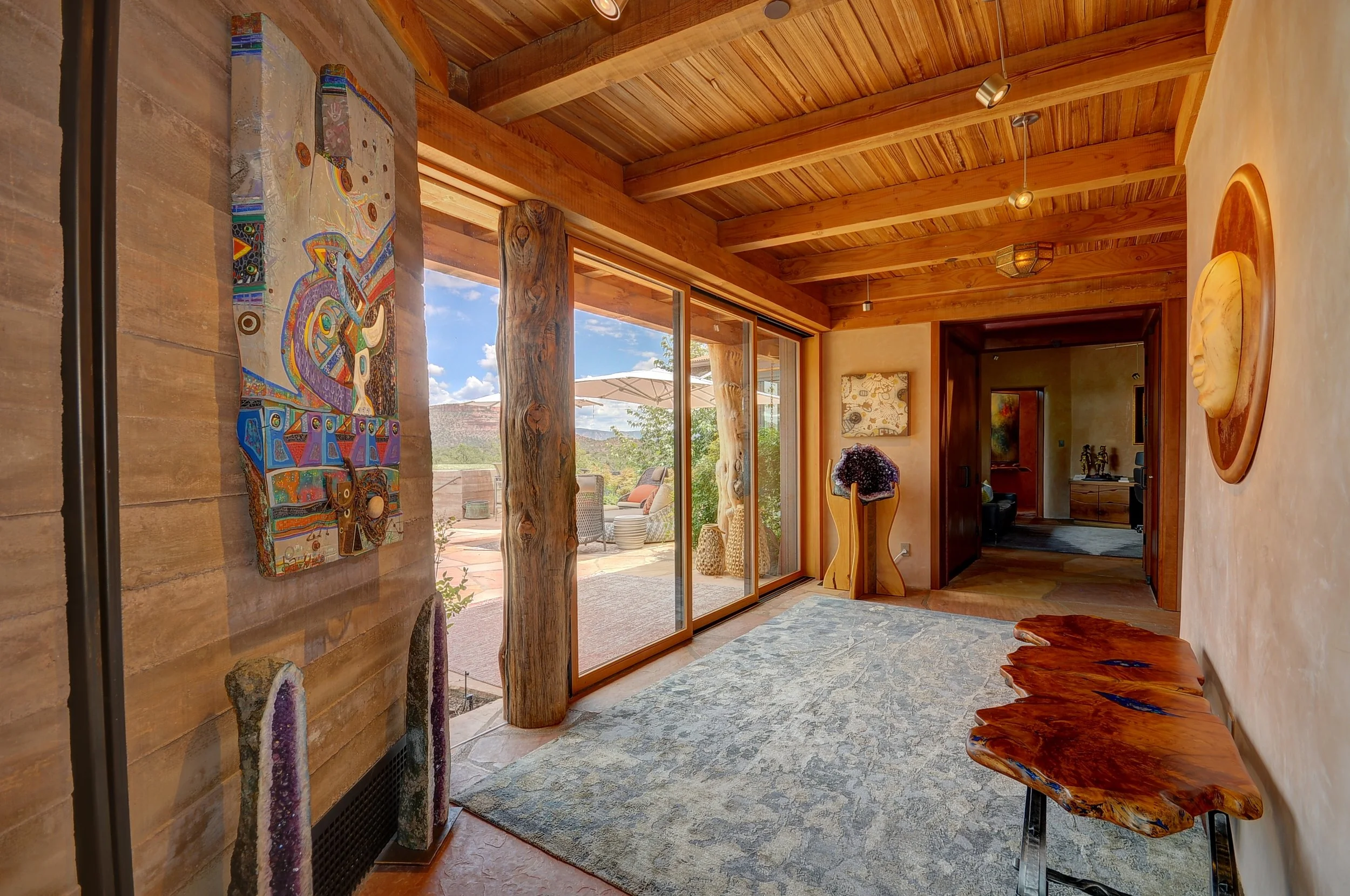 Interior entry hall with wooden ceiling beams, large sliding glass doors leading to an outdoor patio, decorative wall art, and a rustic wooden bench.