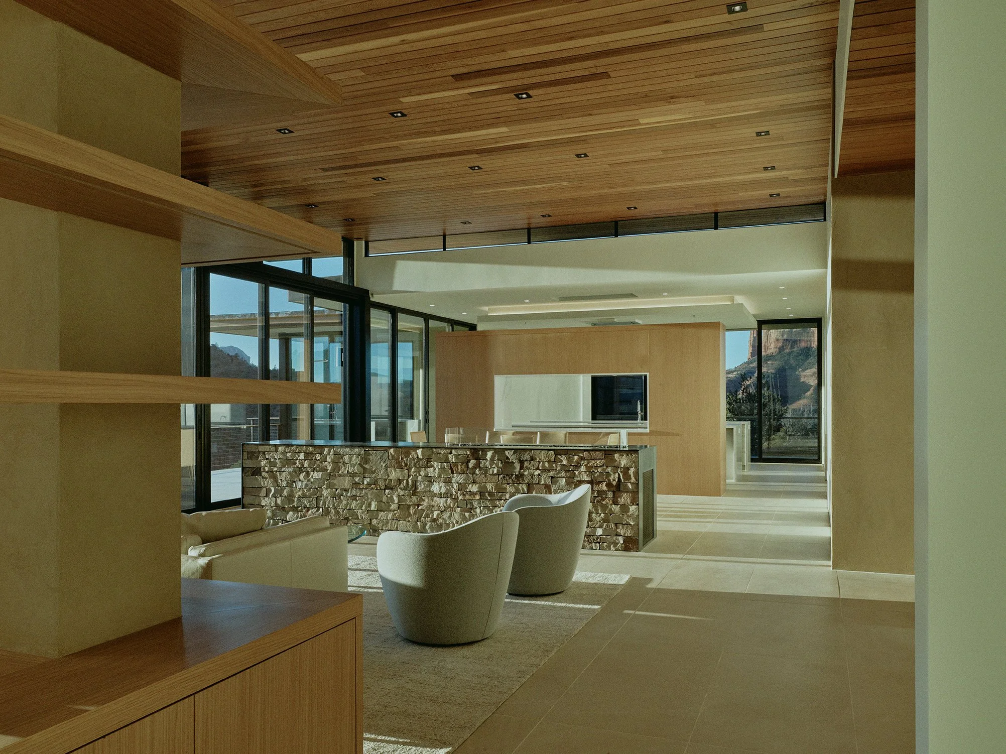 Modern living room with wooden ceiling, glass walls, stone accent wall, beige furniture, and scenic mountain views outside.