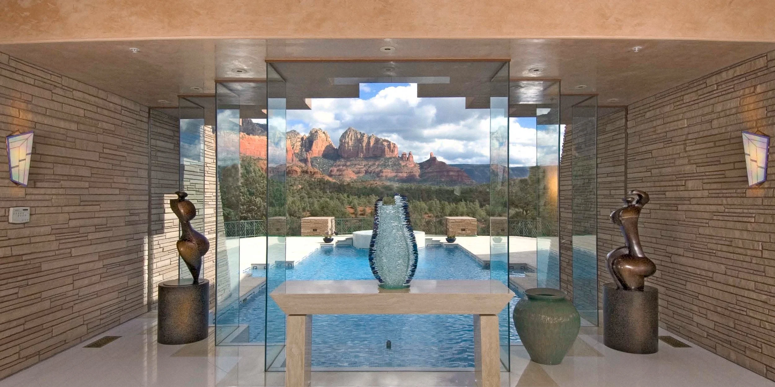 View of a modern interior with glass walls opening to a mountain landscape, a water fountain in a pool, sculptures, and decorative vases.