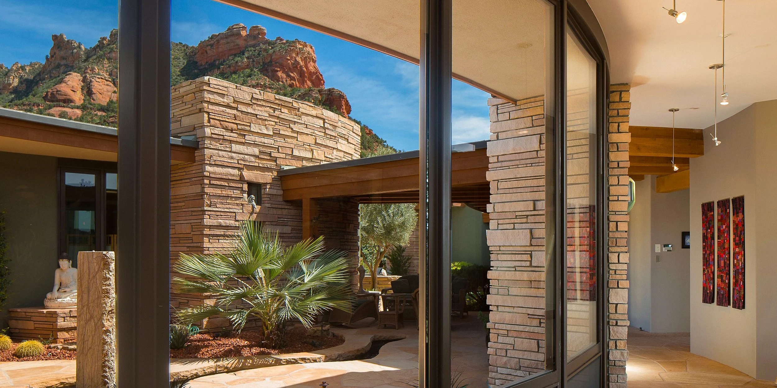 Interior view looking out through large sliding glass doors to an outdoor patio with desert landscaping, a palm tree, and mountains in the background.