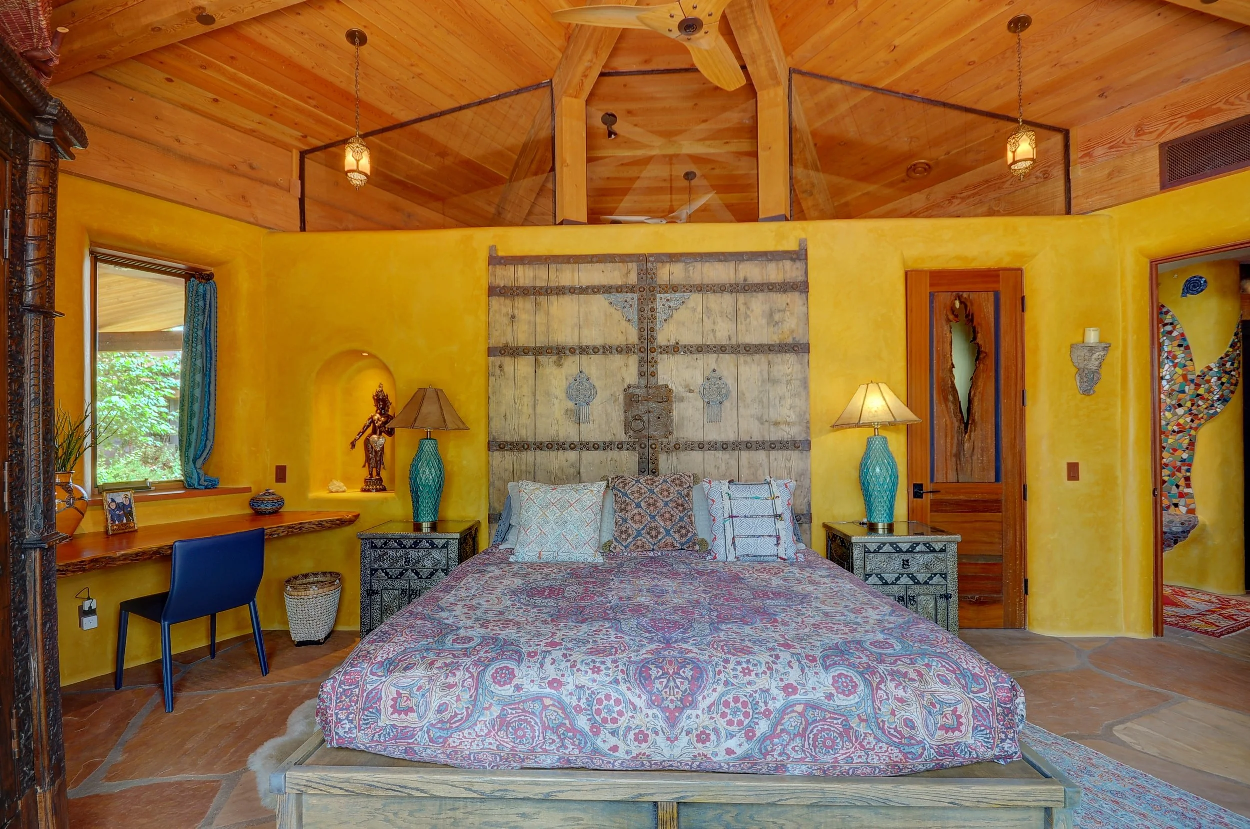 A bedroom with a rustic wooden bed, colorful patterned bedding, yellow walls, turquoise lamps on black patterned nightstands, a small desk with a blue chair near a window with blue curtains, and an arched doorway leading to another room. The ceiling is wooden with hanging lantern lights.