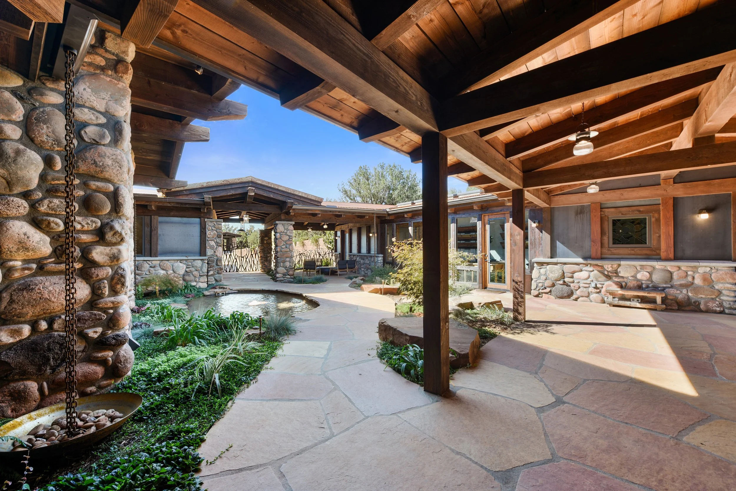 Covered outdoor patio with stone and wood architecture, garden, small pond, and outdoor seating area, under a wooden pergola.
