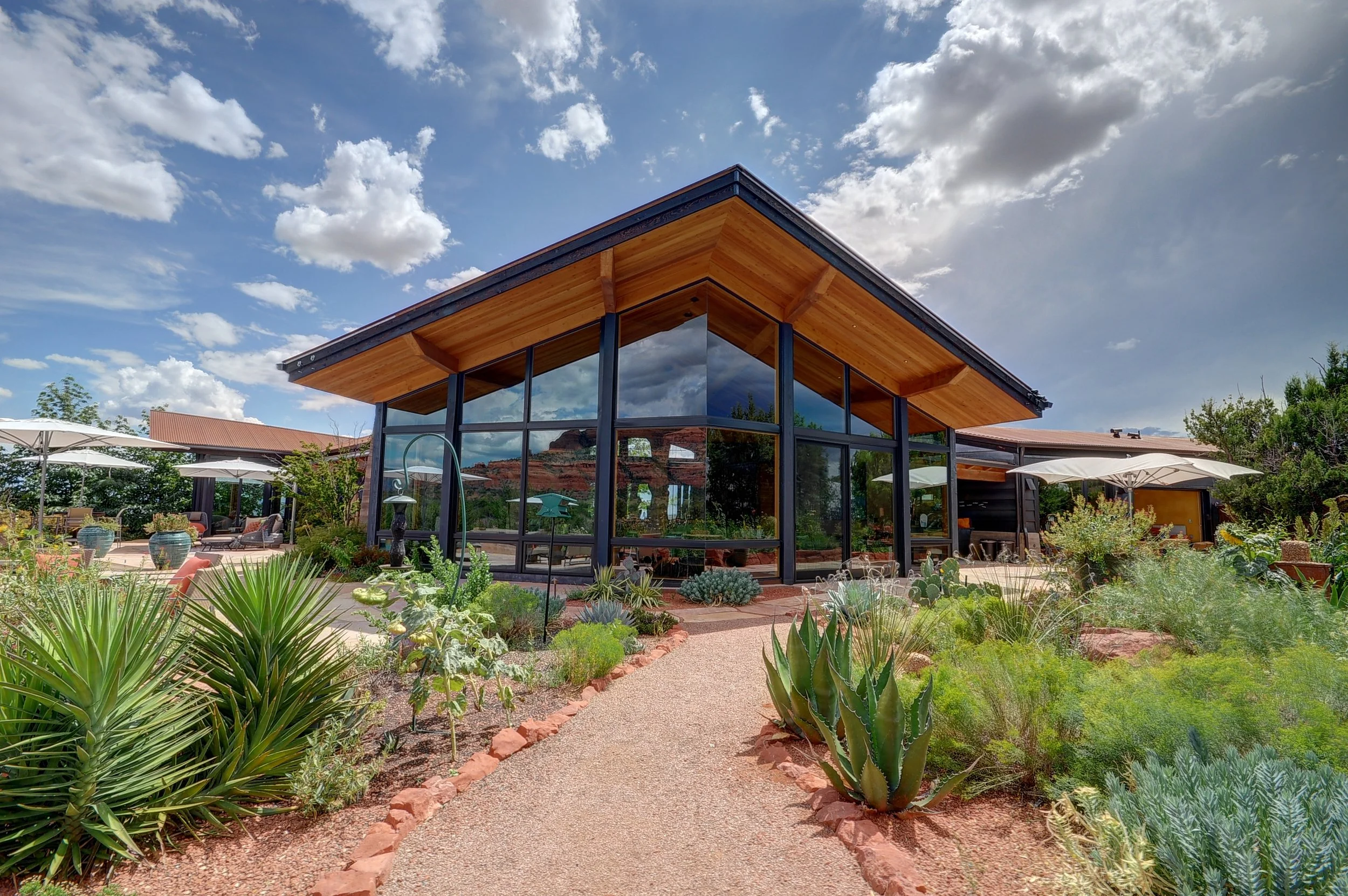 Contemporary southwestern home with large glass windows beneath a flying roof plane, surrounded by desert landscaping, some outdoor seating with umbrellas, under a blue sky with clouds.