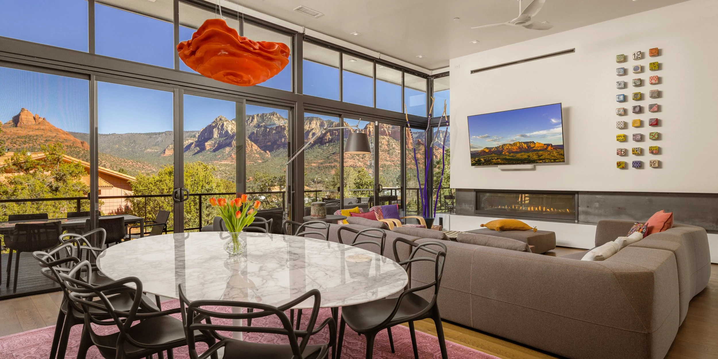 Modern living room with a large wall of windows showing mountain views, a round marble dining table with a vase of tulips, a sectional sofa with colorful cushions, and a fireplace below a mounted TV.